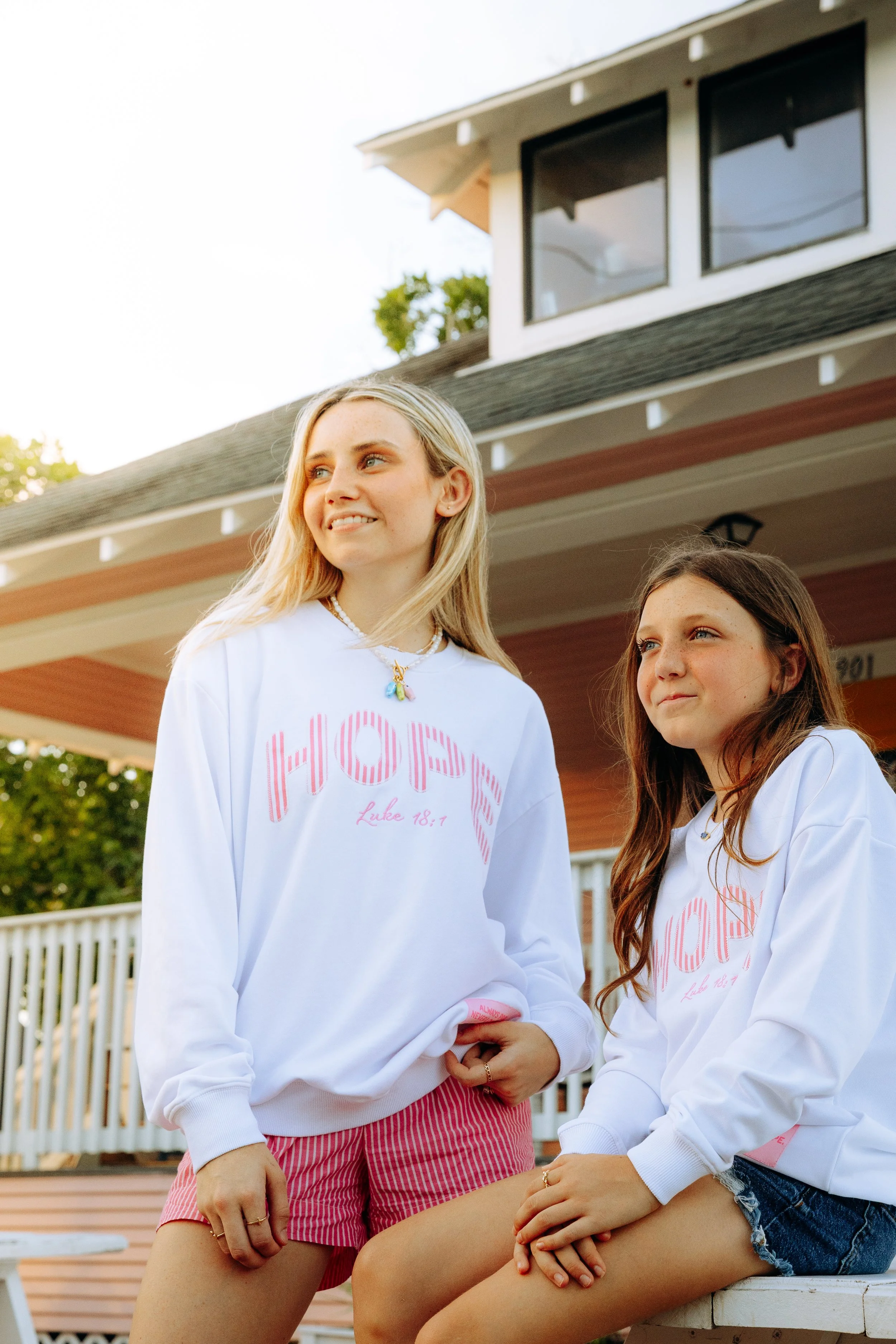 Two young women sitting outdoors in front of a house, wearing white sweatshirts with pink message and red and pink striped shorts. One is sitting on a bench, while the other stands nearby. The background shows a white fence and a house with large win