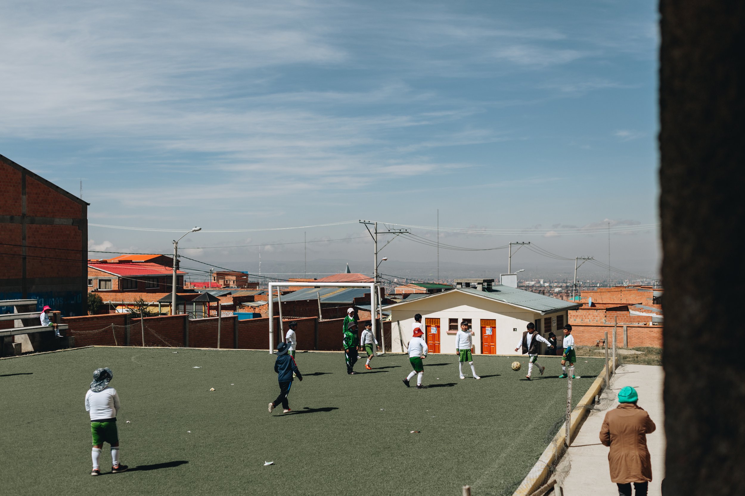 Children playing soccer on a field in a neighborhood with houses and utility poles in the background under a blue sky.