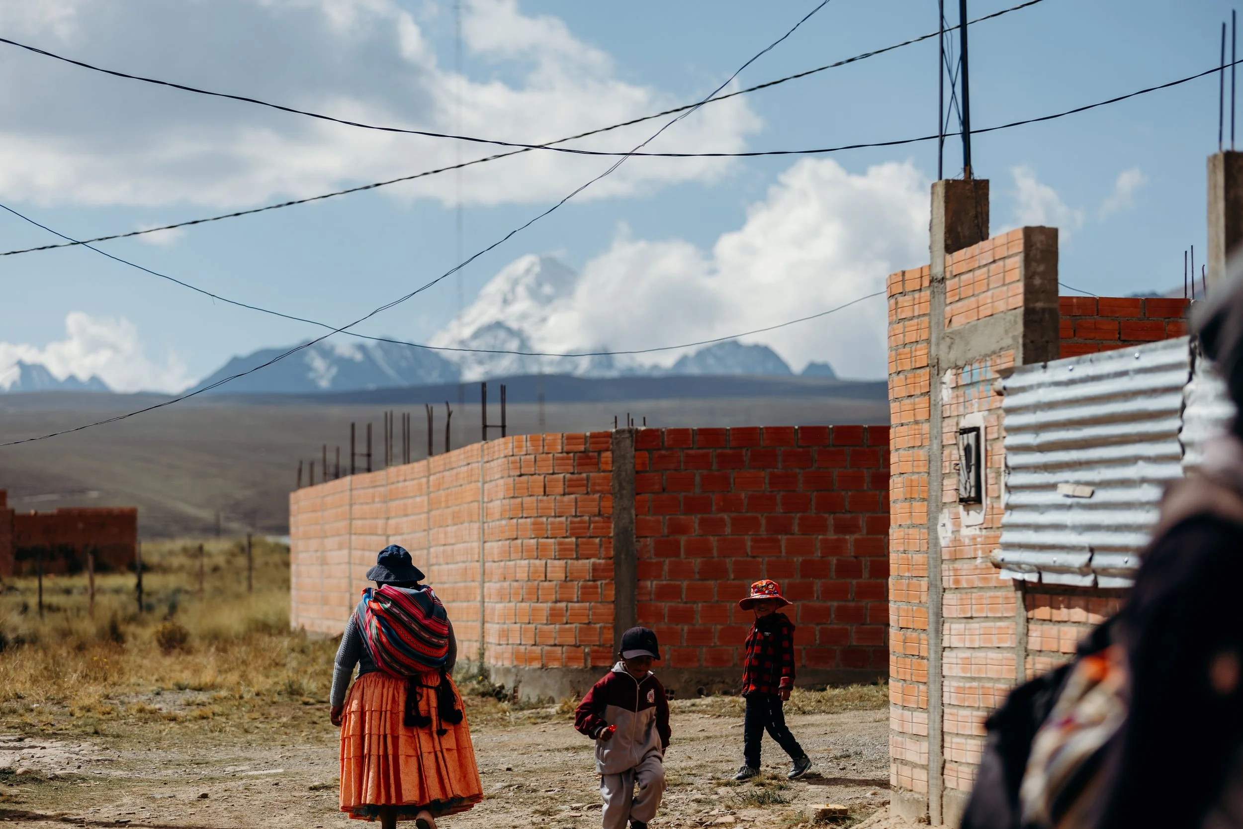 Children playing in a rural village with brick and corrugated metal buildings, mountains in the background, and blue sky with clouds.