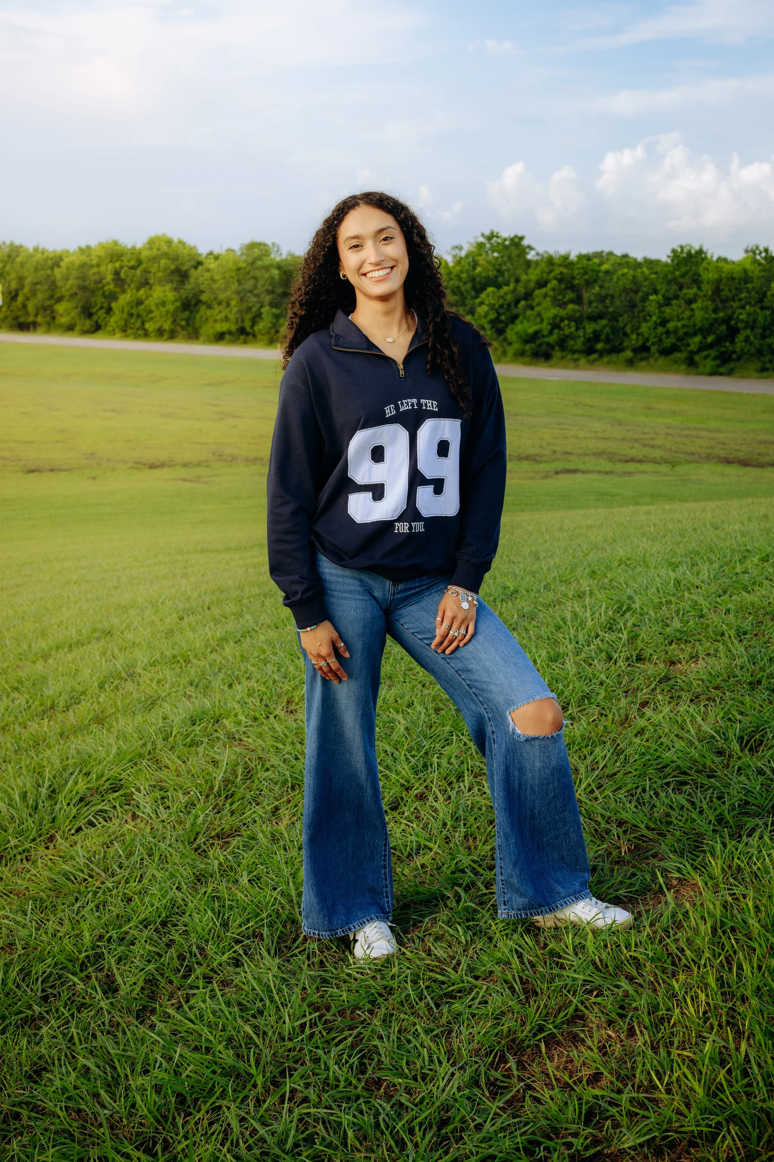 A young woman with curly hair standing on a grassy field, smiling, wearing a navy blue sweatshirt with the number 99 and jeans with a ripped knee.