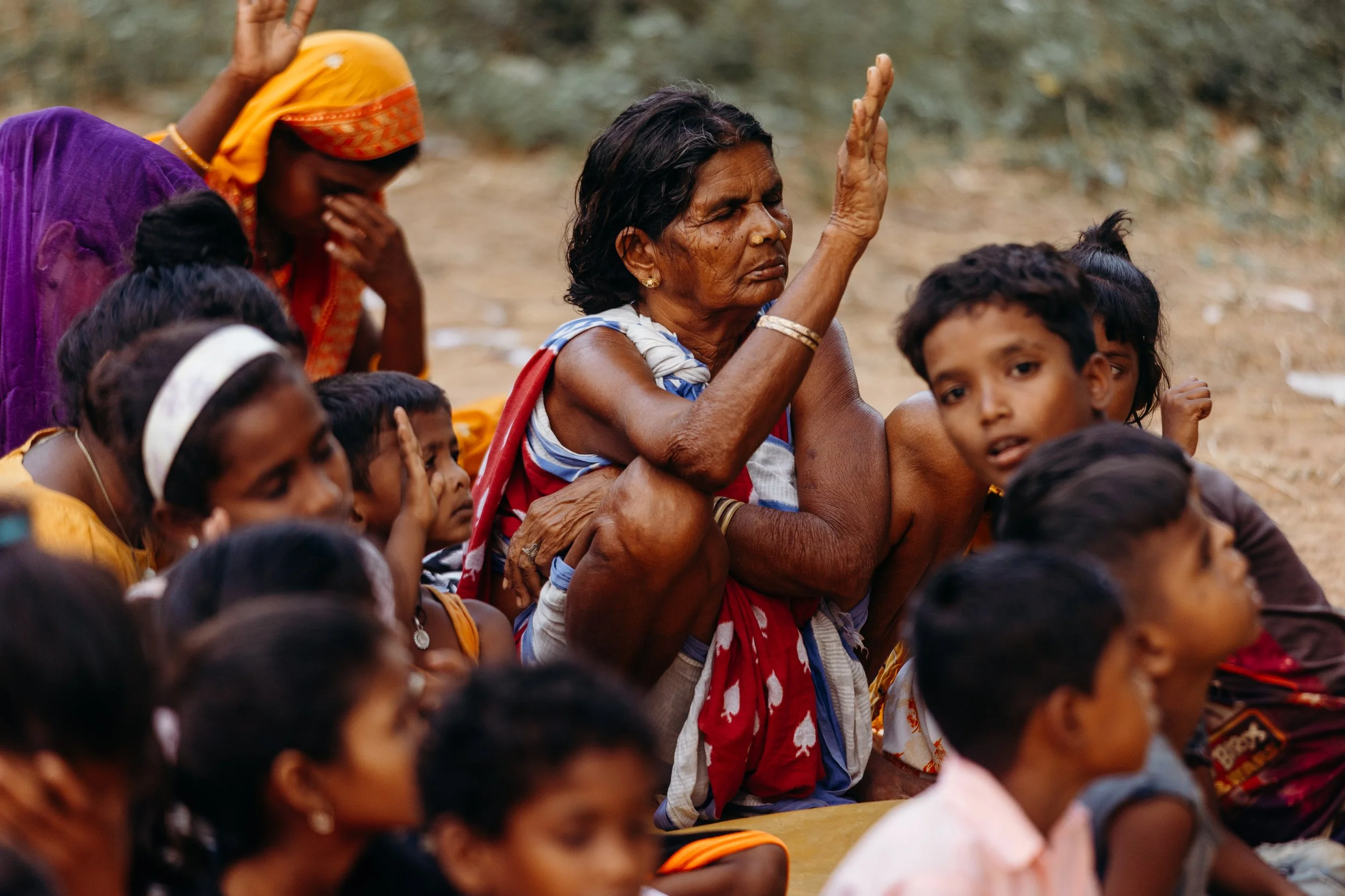 An elderly woman with dark hair tied back, wearing a red and white sari, raises her right hand while sitting on the ground surrounded by children. The children are sitting close, some with expressions of concern, sadness, or curiosity, in an outdoor 