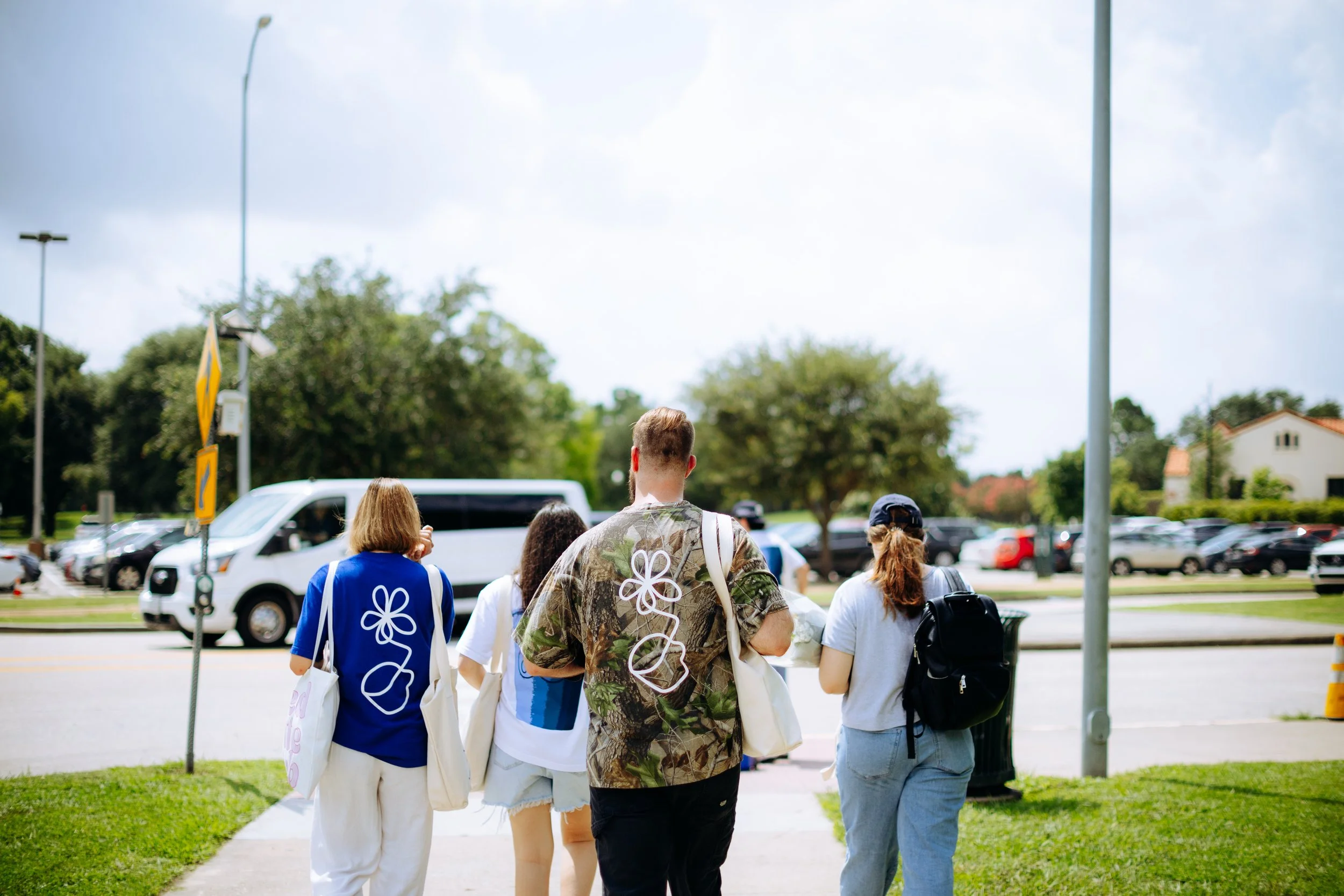 Group of five people walking outdoors near a parking lot, with trees and a cloudy sky in the background.