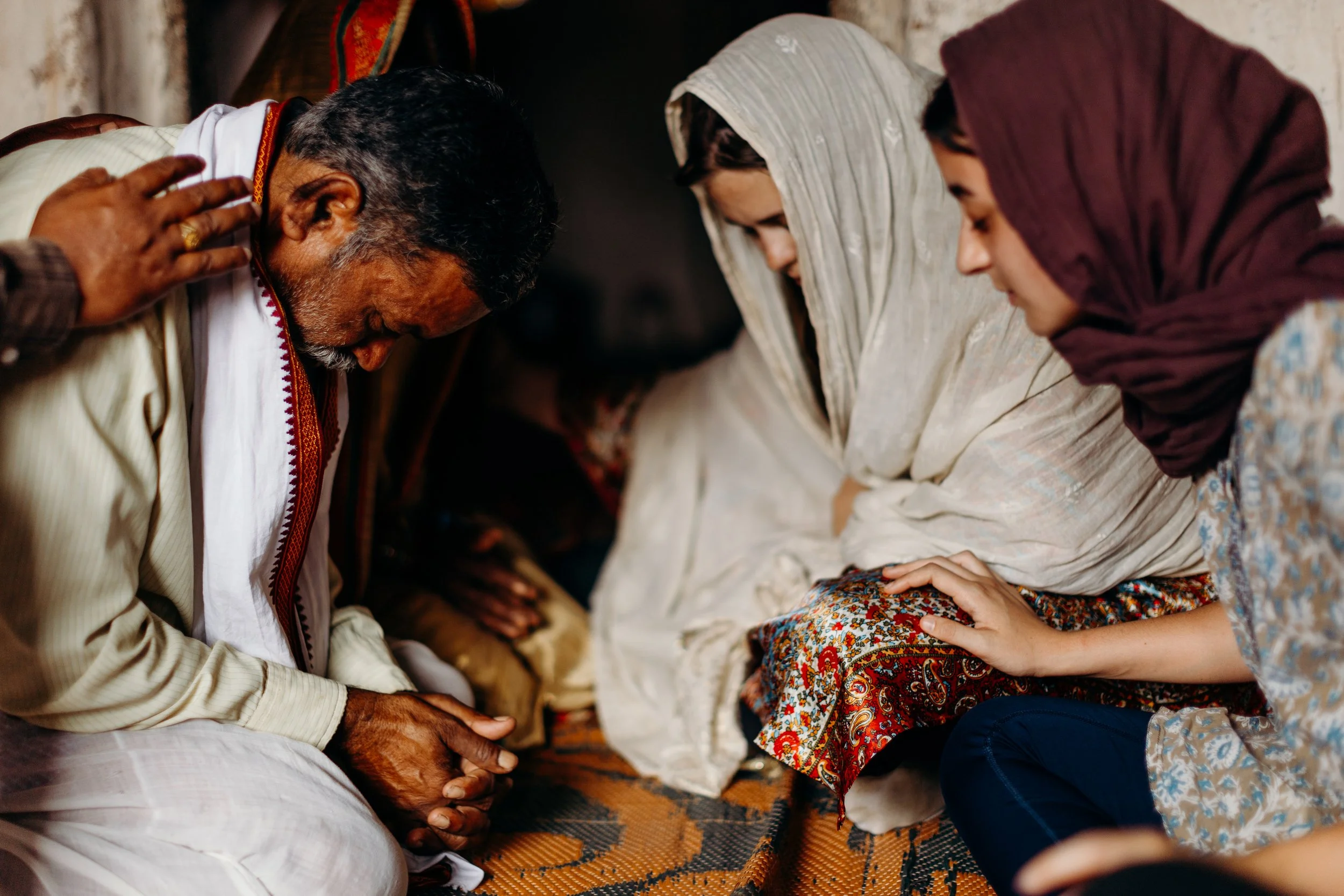 Group of people praying together with their heads bowed and hands placed on their knees, sitting on a patterned rug.