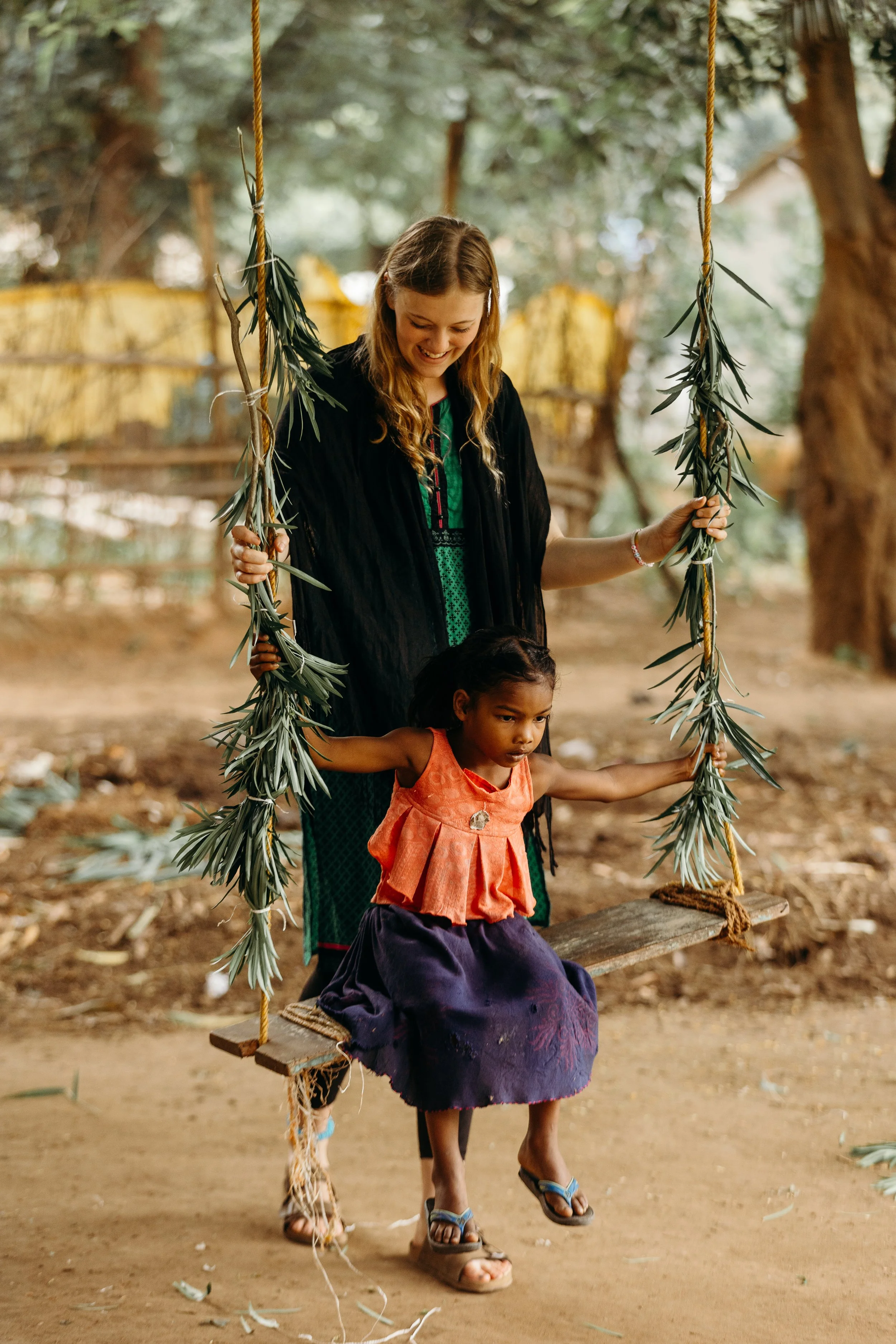 A woman with long, wavy hair in a black cardigan smiling as she pushes a young girl on a wooden swing decorated with green leaves, set outdoors with trees in the background.