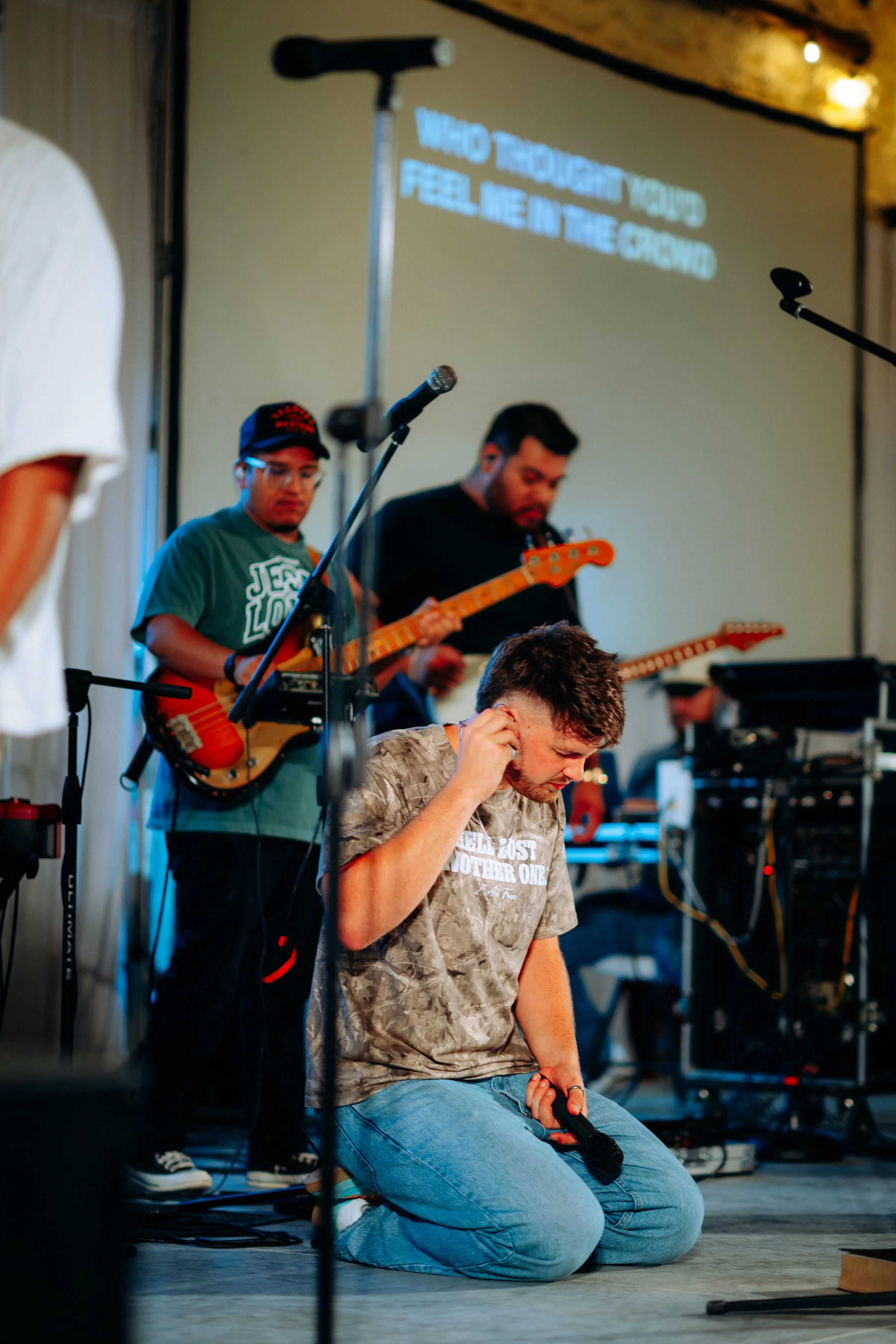 A group of musicians performing on stage, including a kneeling man holding a microphone and a man playing guitar behind him, with lyrics projected on a screen in the background.