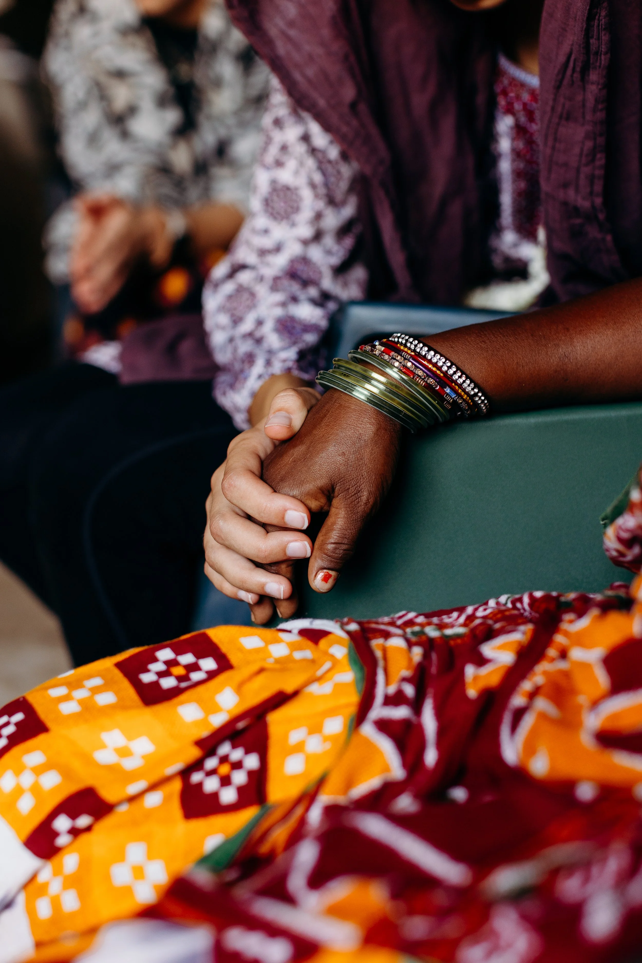 Two people holding hands, one with dark skin wearing multiple colorful bangles, and the other with lighter skin, surrounded by patterned clothing and a background with a person in a patterned dress and what appears to be a dog.