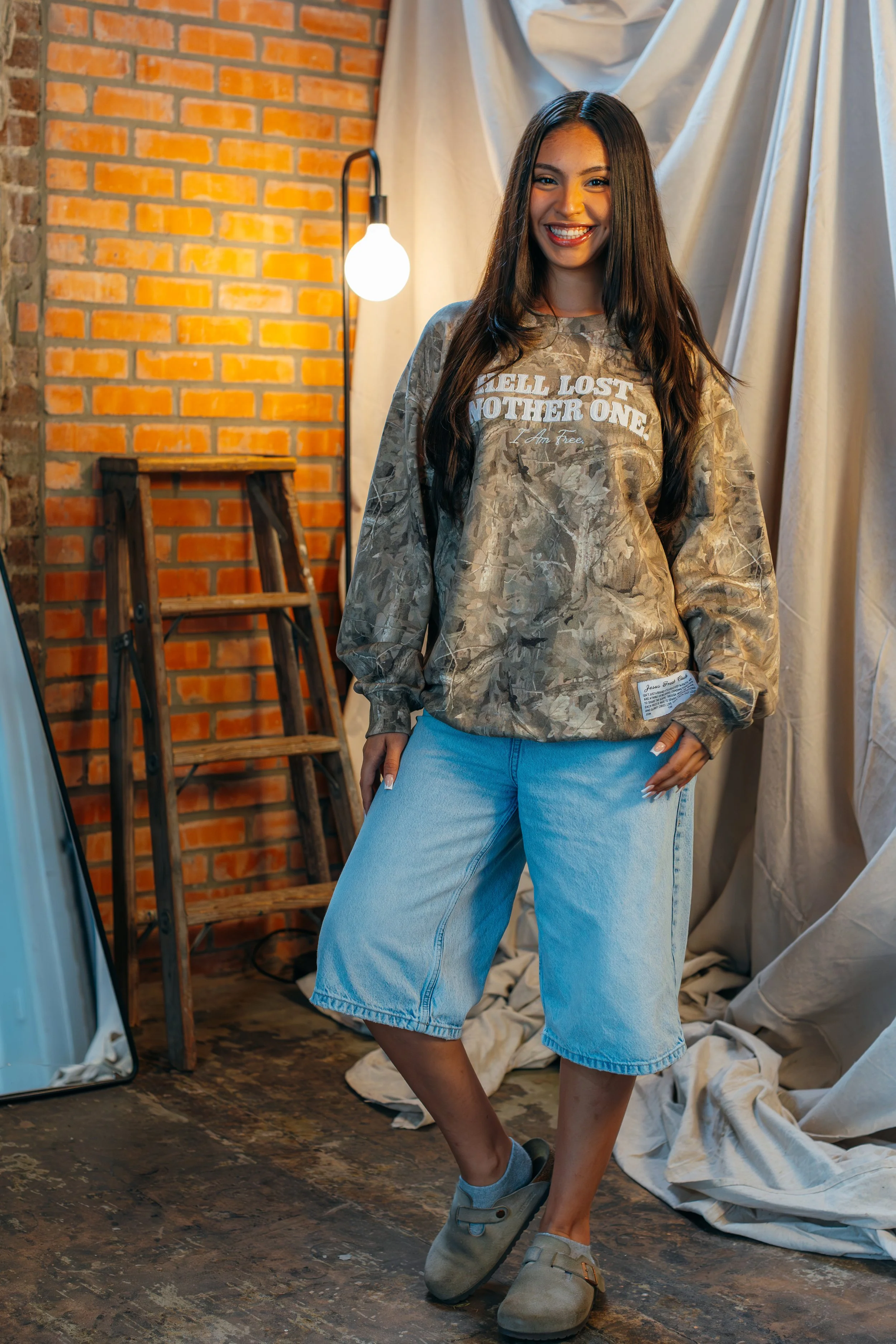 A young woman with long dark hair and a bright smile stands indoors in front of a brick wall and a curtain. She is wearing a camouflage sweatshirt with white text and light blue knee-length denim shorts, along with gray shoes and socks. She poses wit