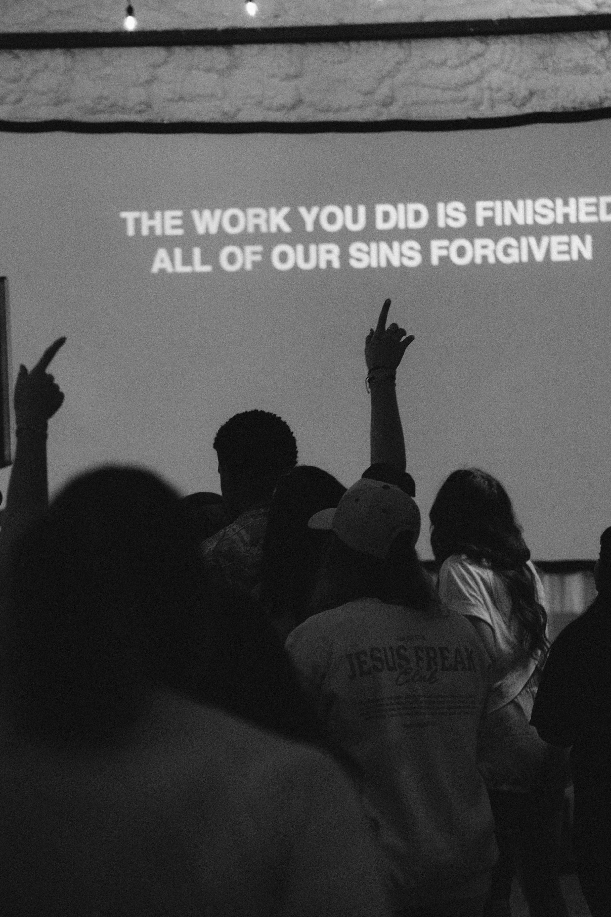 A group of people in a dark room watching a presentation projected on a screen, with two individuals raising their hands. The slide on the screen reads, 'THE WORK YOU DID IS FINISHED, ALL OF OUR SINS FORGIVEN.'