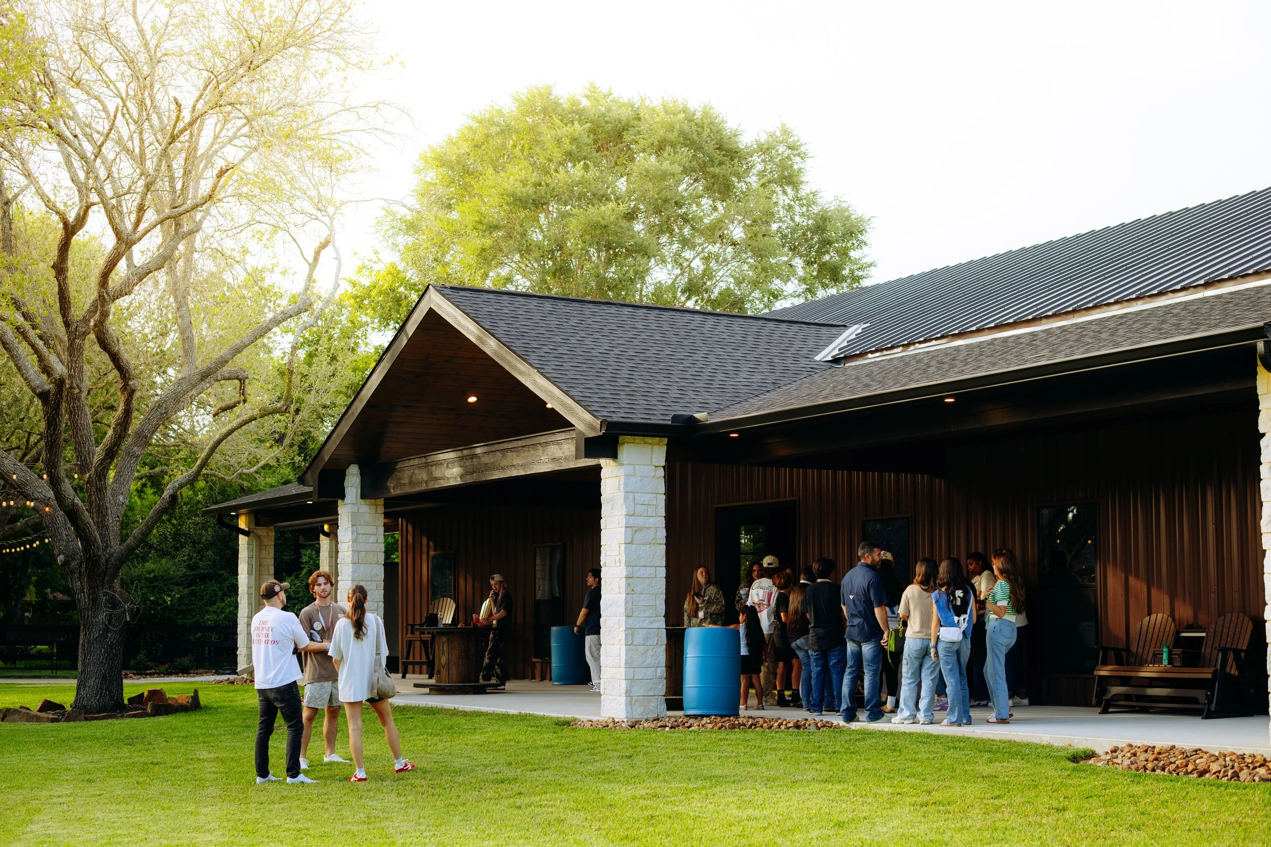 People gathered outside a modern building with a porch, lined up and socializing on a grassy area under a tree.