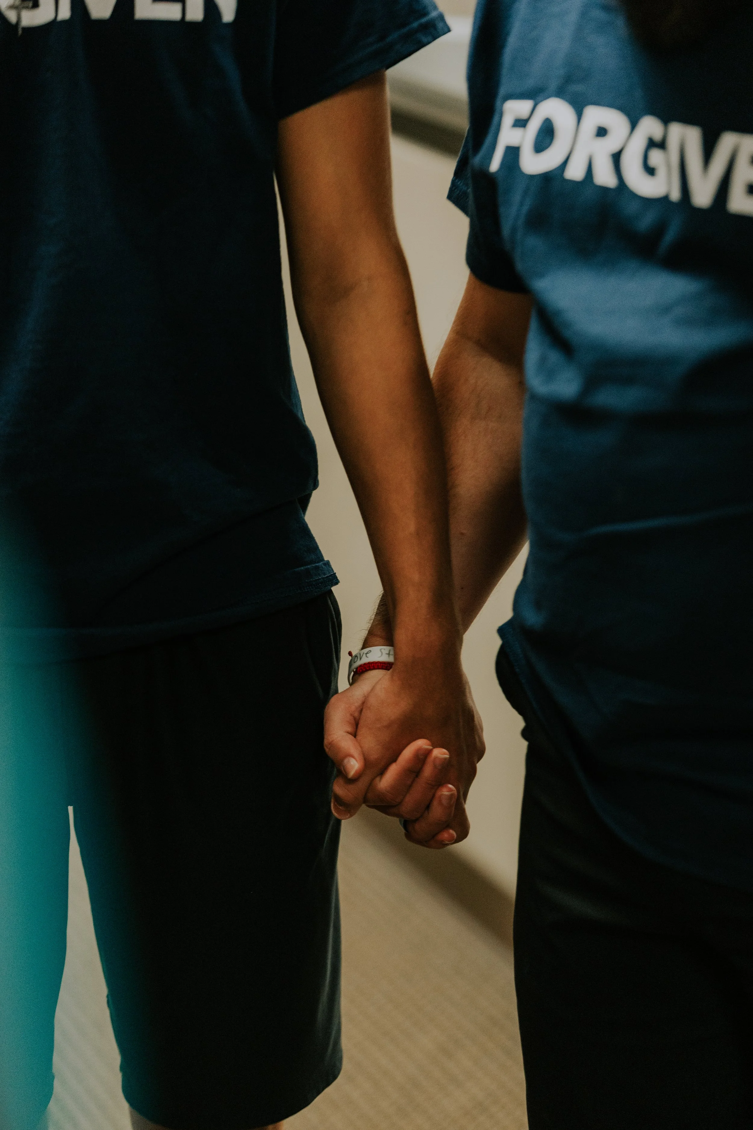 Two people holding hands, wearing navy T-shirts with one partially showing the word 'FORGIVE' and a wristband with a red bracelet.