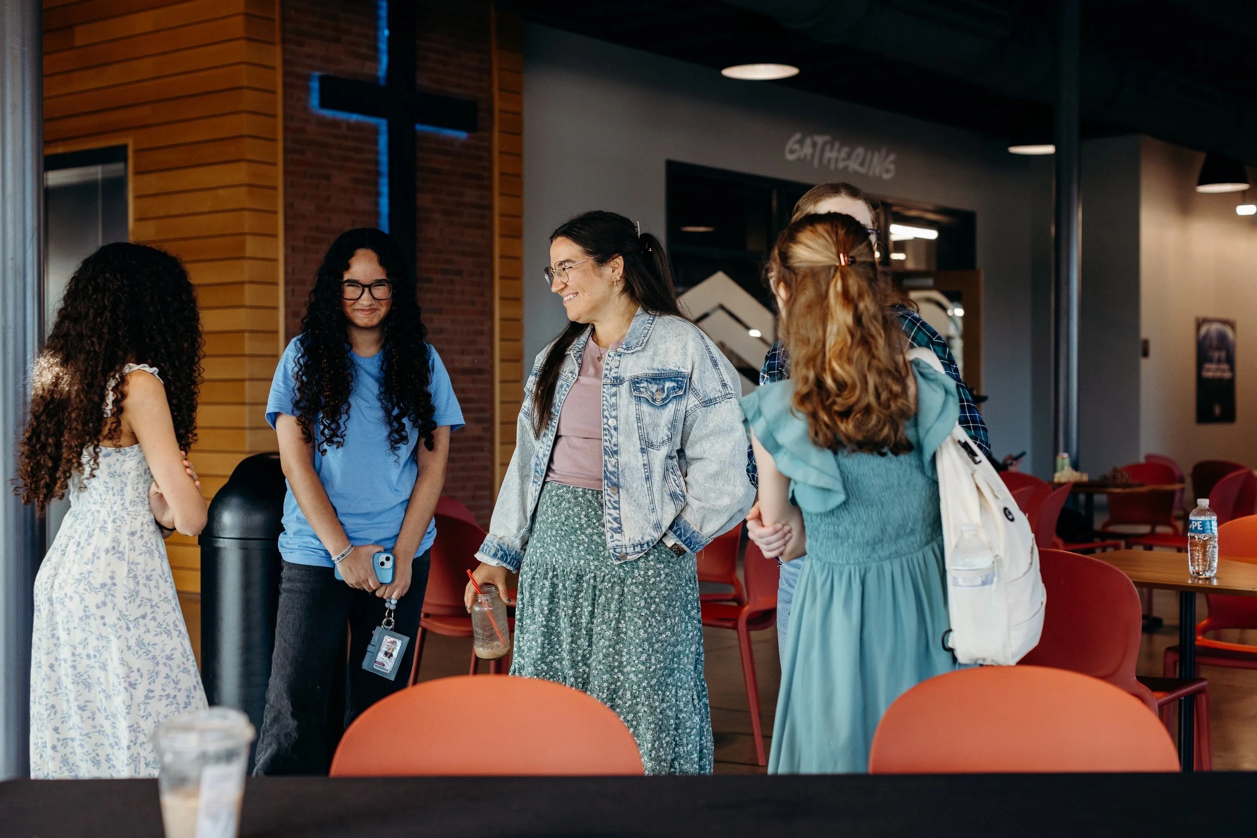 A group of five young women engaged in conversation in a modern indoor space, with a large cross illuminated on the brick wall in the background.