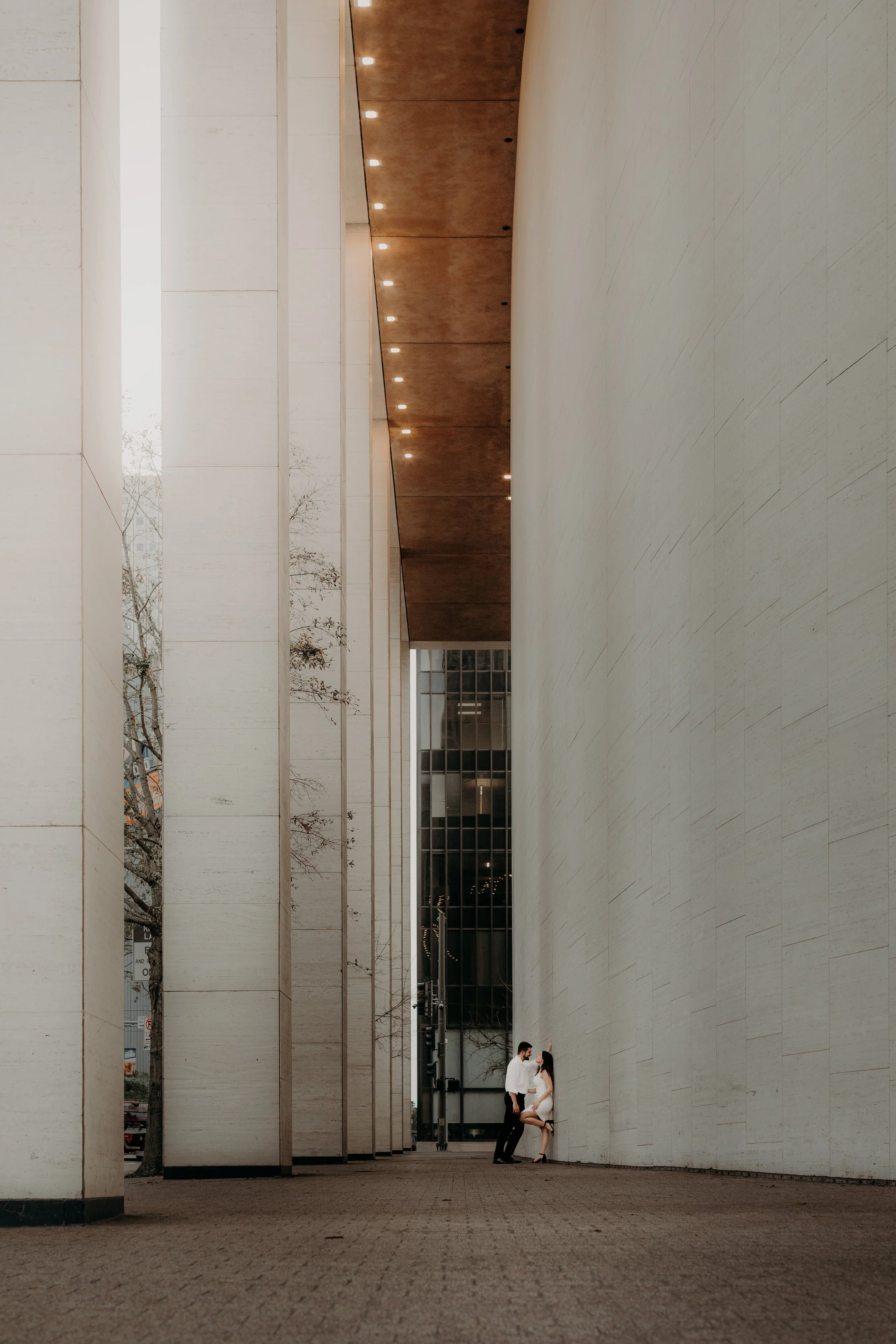 A man and woman in formal attire standing against a large, modern architectural wall outside a building.