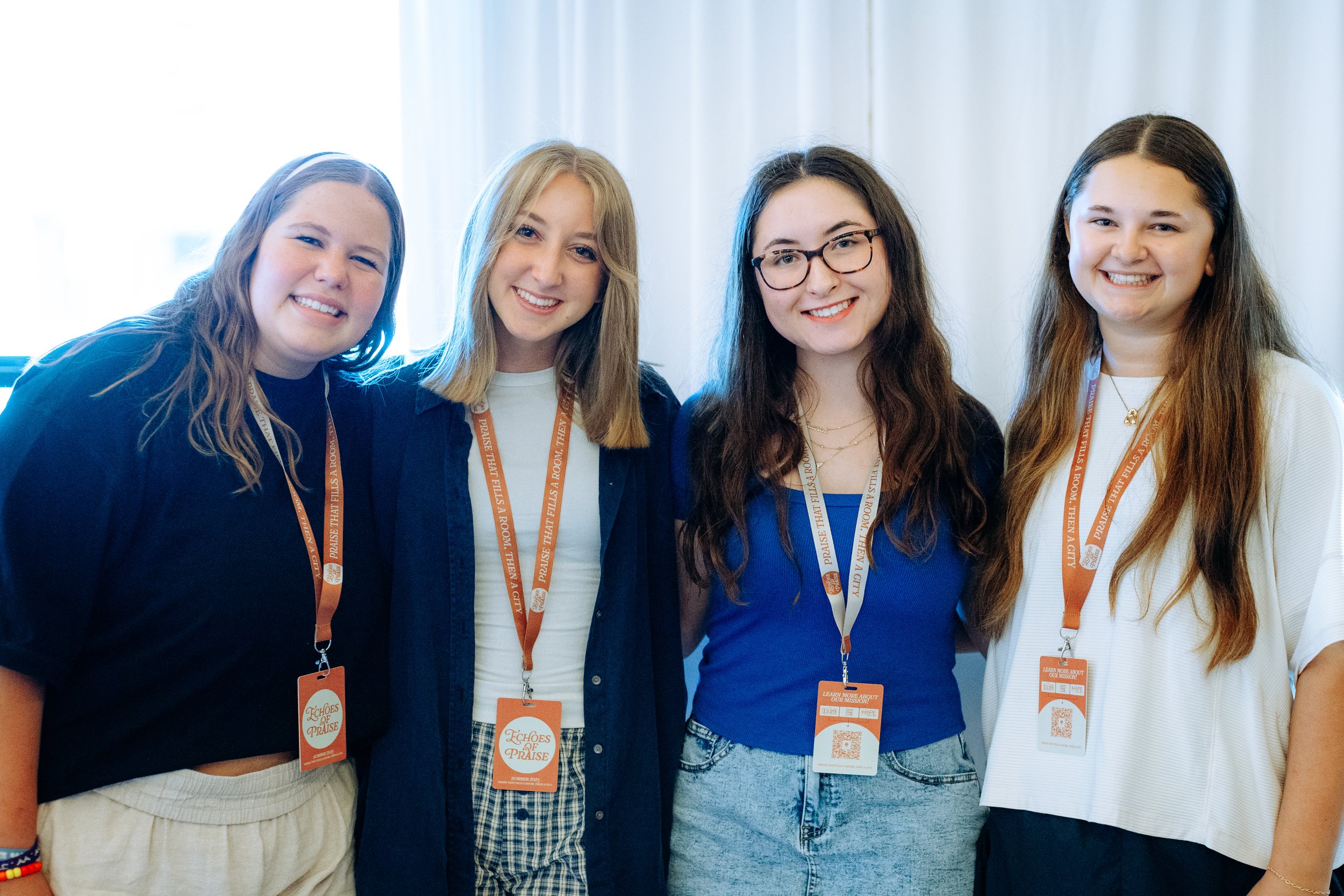 Four young women standing side by side smiling at the camera, each wearing a lanyard and badge, and casual clothing.