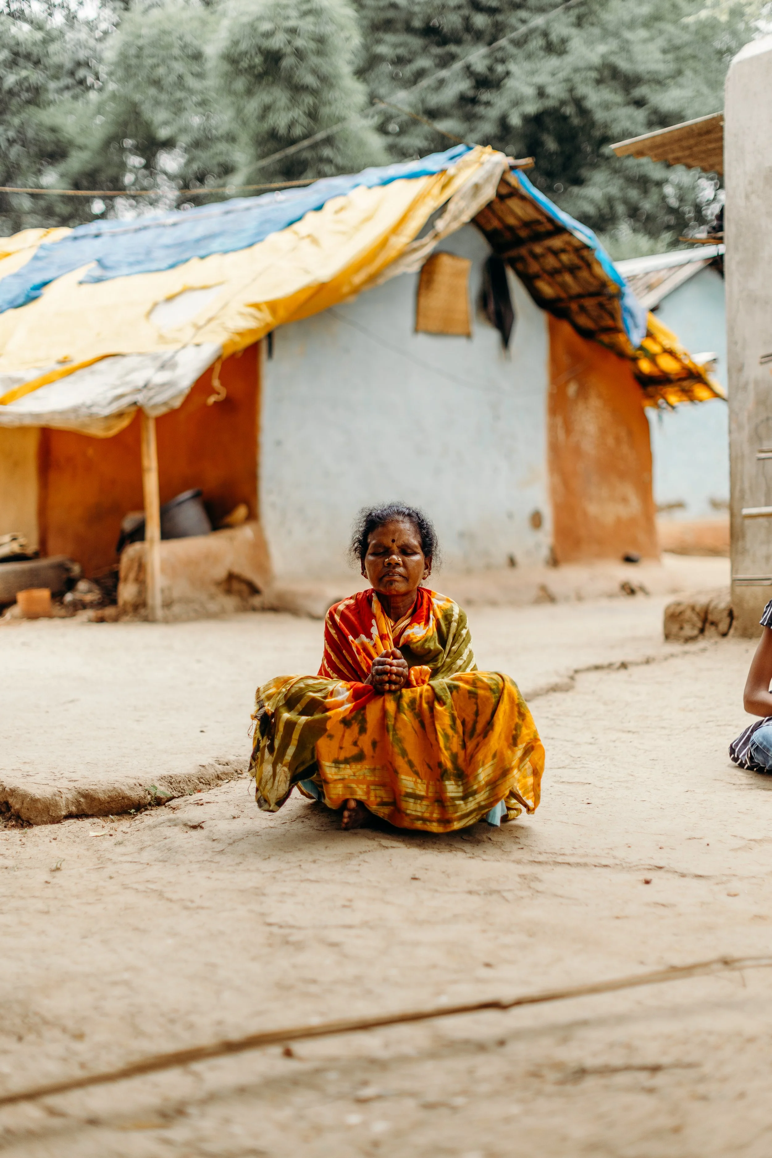A woman sitting cross-legged and praying outdoors in front of a modest house with a blue, yellow, and brown tarp roof in a rural setting.