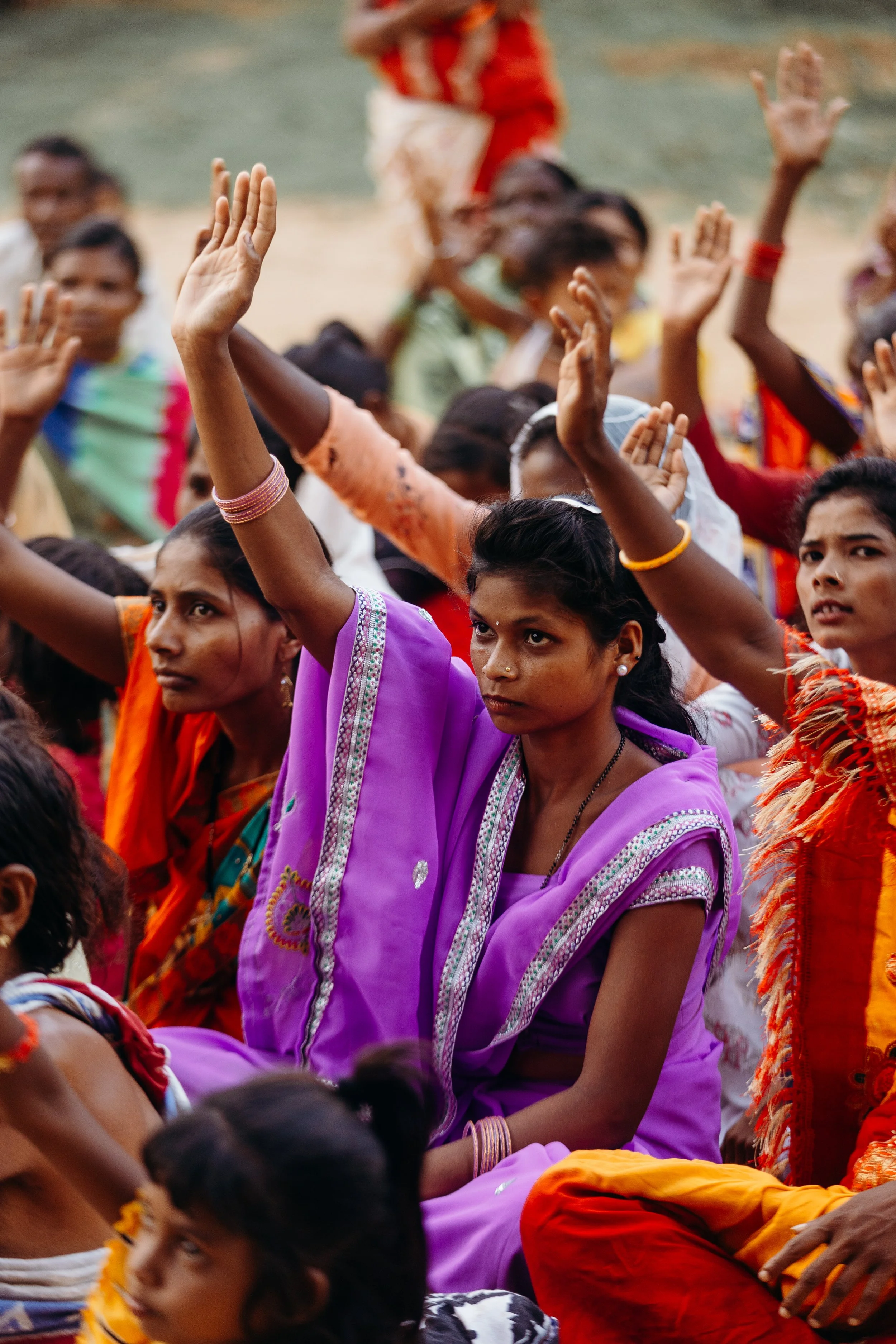 Group of women and children sitting outdoors, raising their hands, some wearing colorful traditional clothing.