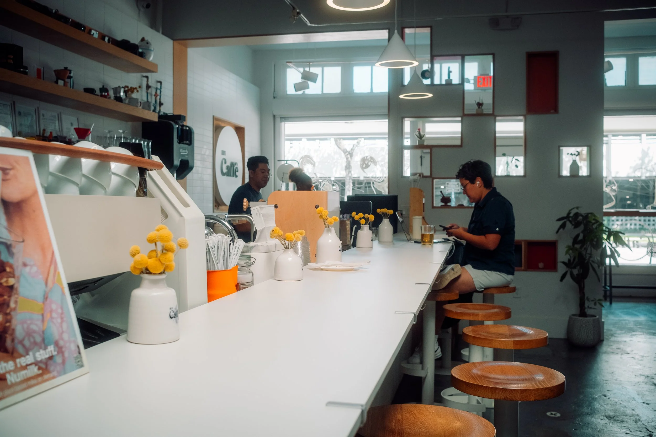 Inside a modern cafe with several customers sitting at a white counter, decorated with white vases of yellow flowers, with shelves and large windows in the background.