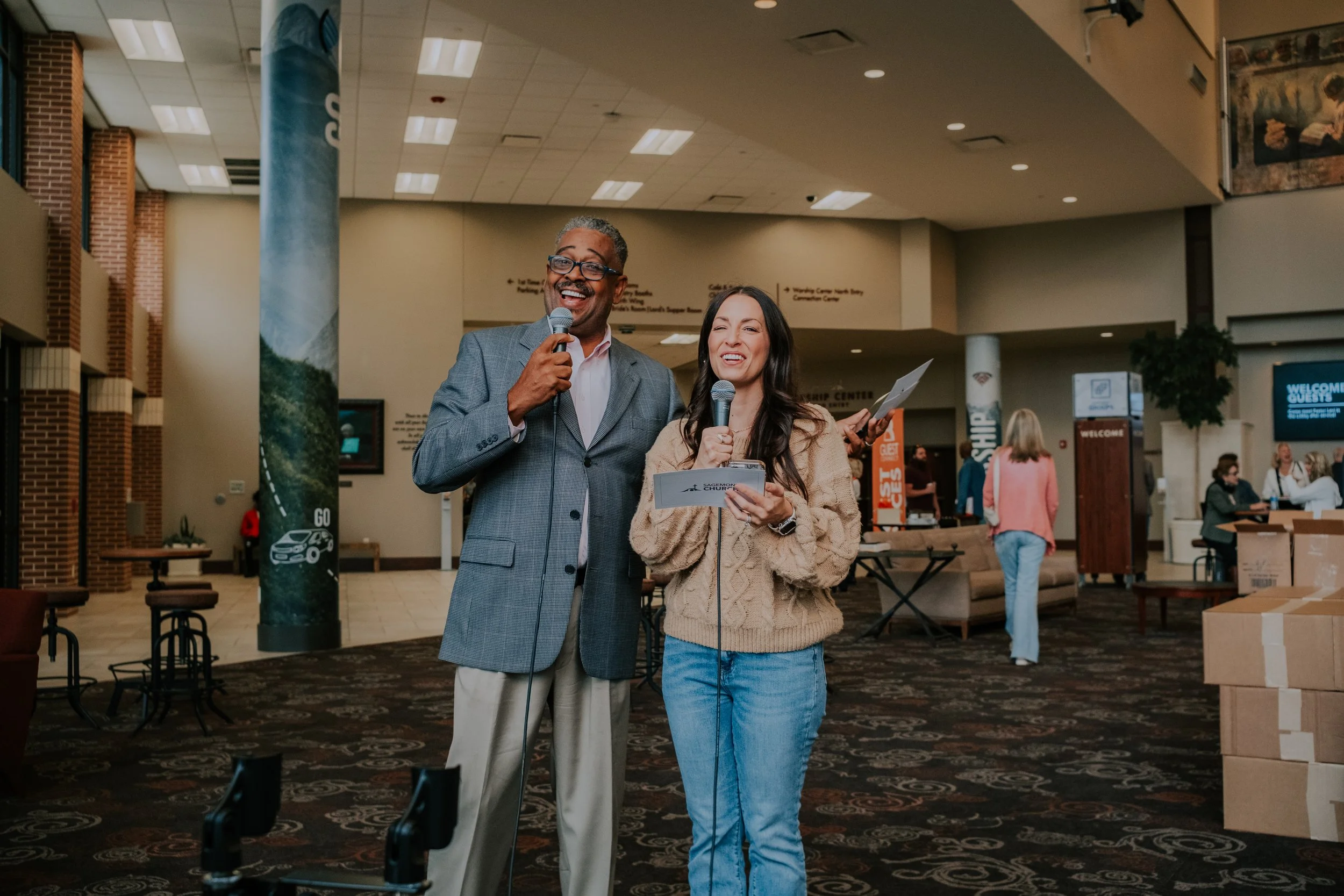 A man and a woman standing indoors, smiling, holding microphones and papers, likely hosting or speaking at an event. The man wears a gray blazer, the woman a beige sweater, and there are other people and event materials in the background.