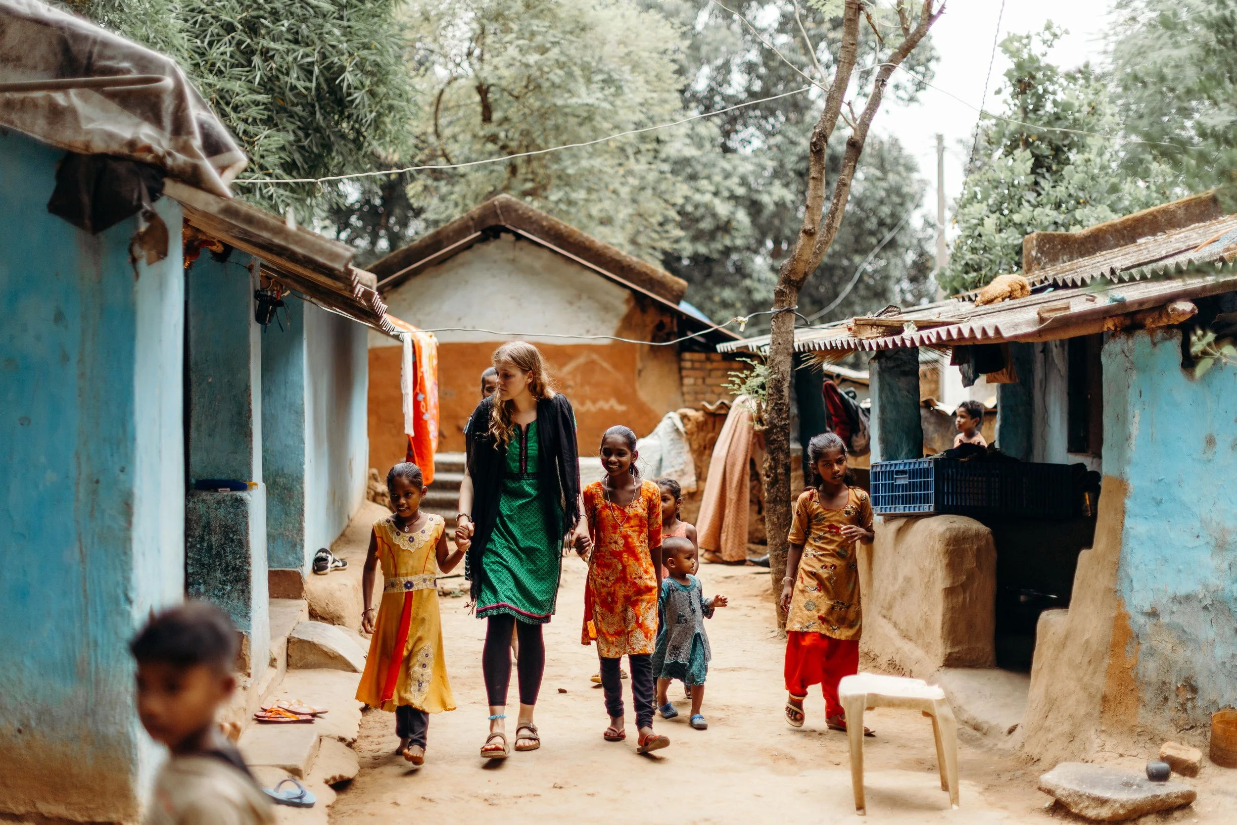 Women and children walking through a rustic village with mud and stone houses, some covered with tin roofs, surrounded by trees and outdoor clotheslines.