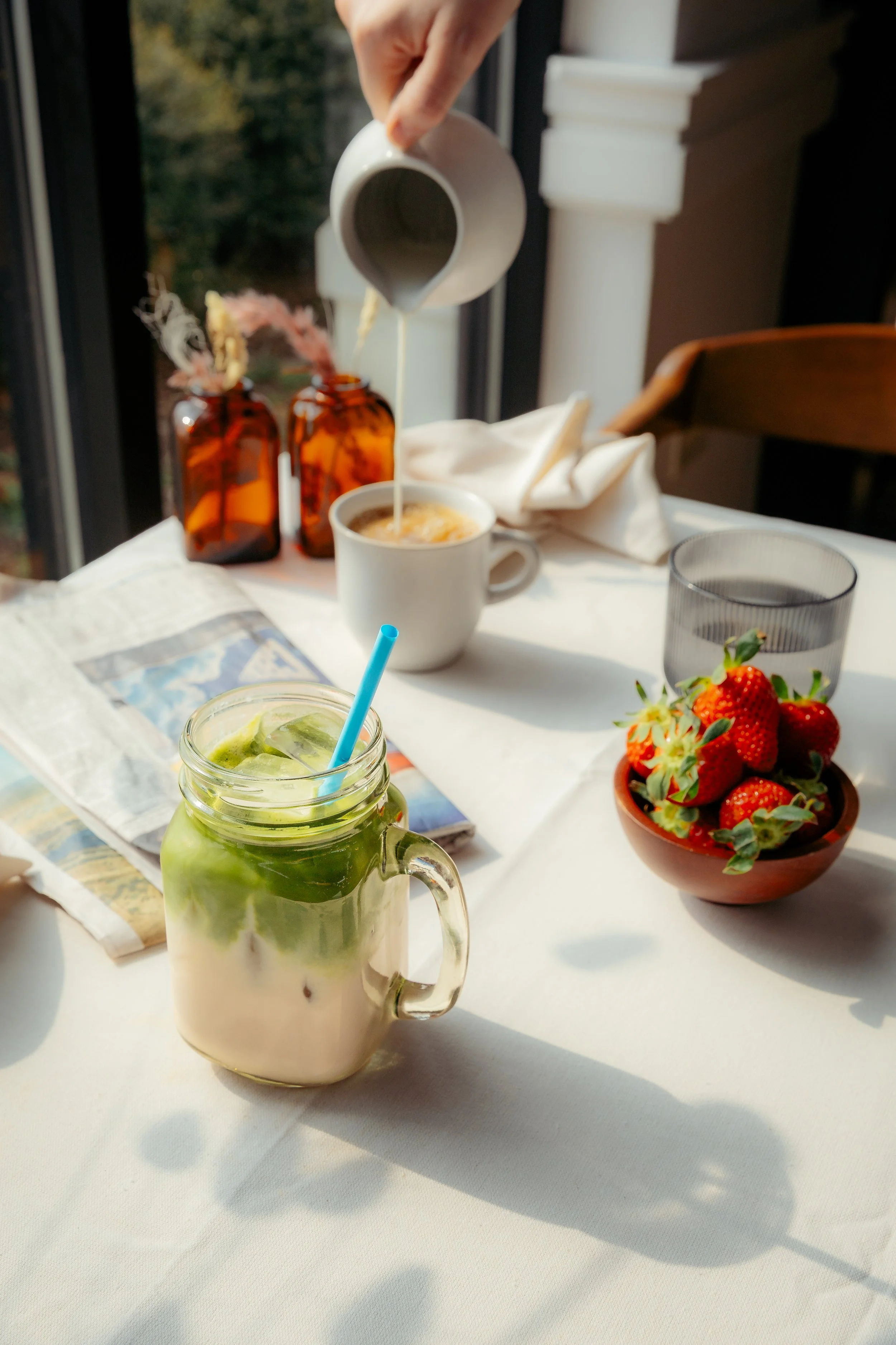 A table set with drinks and food. In the foreground, a glass jar with a handle contains a kiwi and coconut smoothie with a blue straw. Behind it, a white mug has a frothy coffee or latte being poured from a small white pitcher, with steam rising from the beverage. To the right, a bowl with strawberries is visible. In the background, two amber glass bottles hold dried flowers, with a window showing trees outside. There is also a folded napkin and a map on the table.