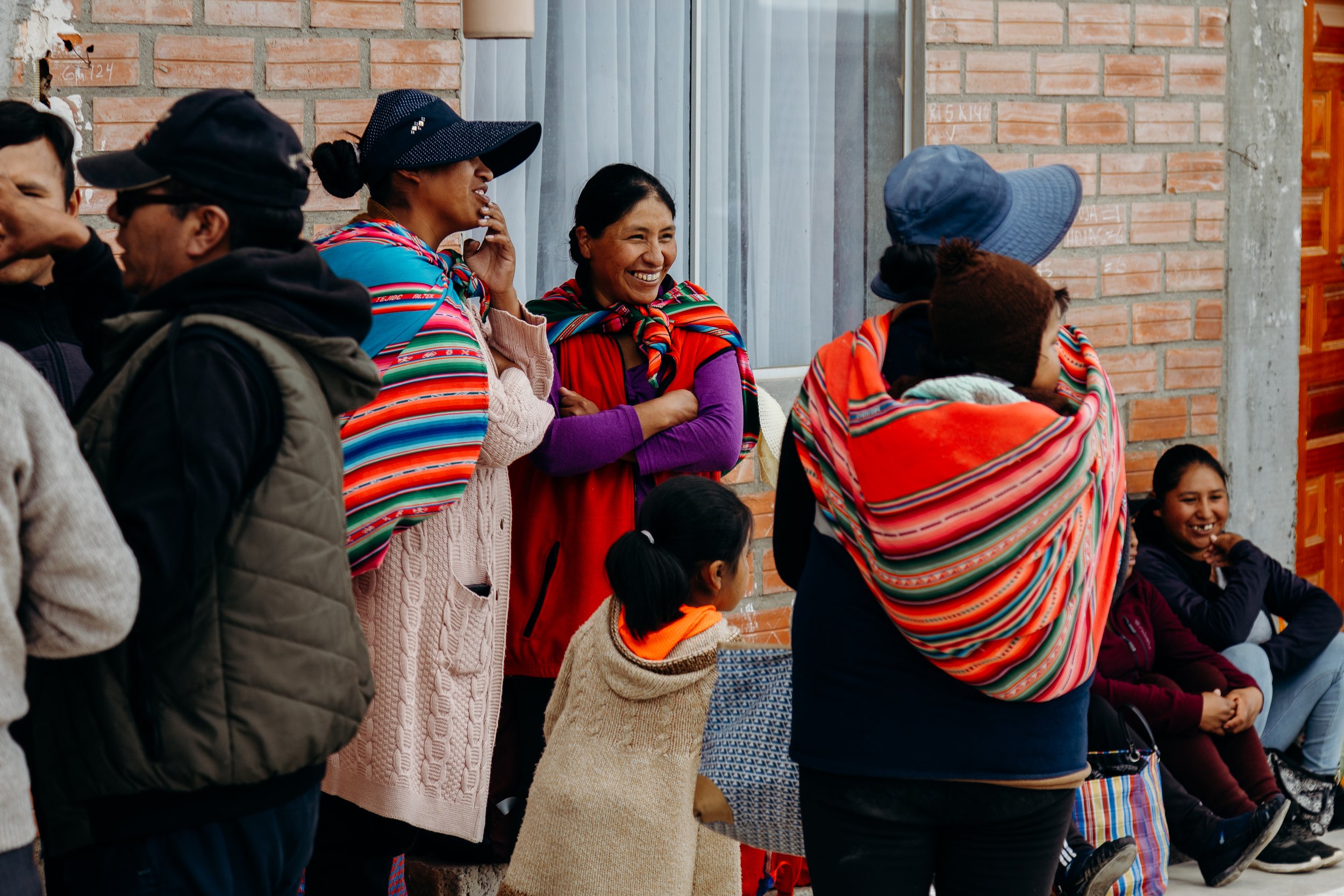 Group of people, including women and children, wearing traditional colorful, striped clothing and ponchos, gathered outdoors against a brick wall, smiling and interacting.