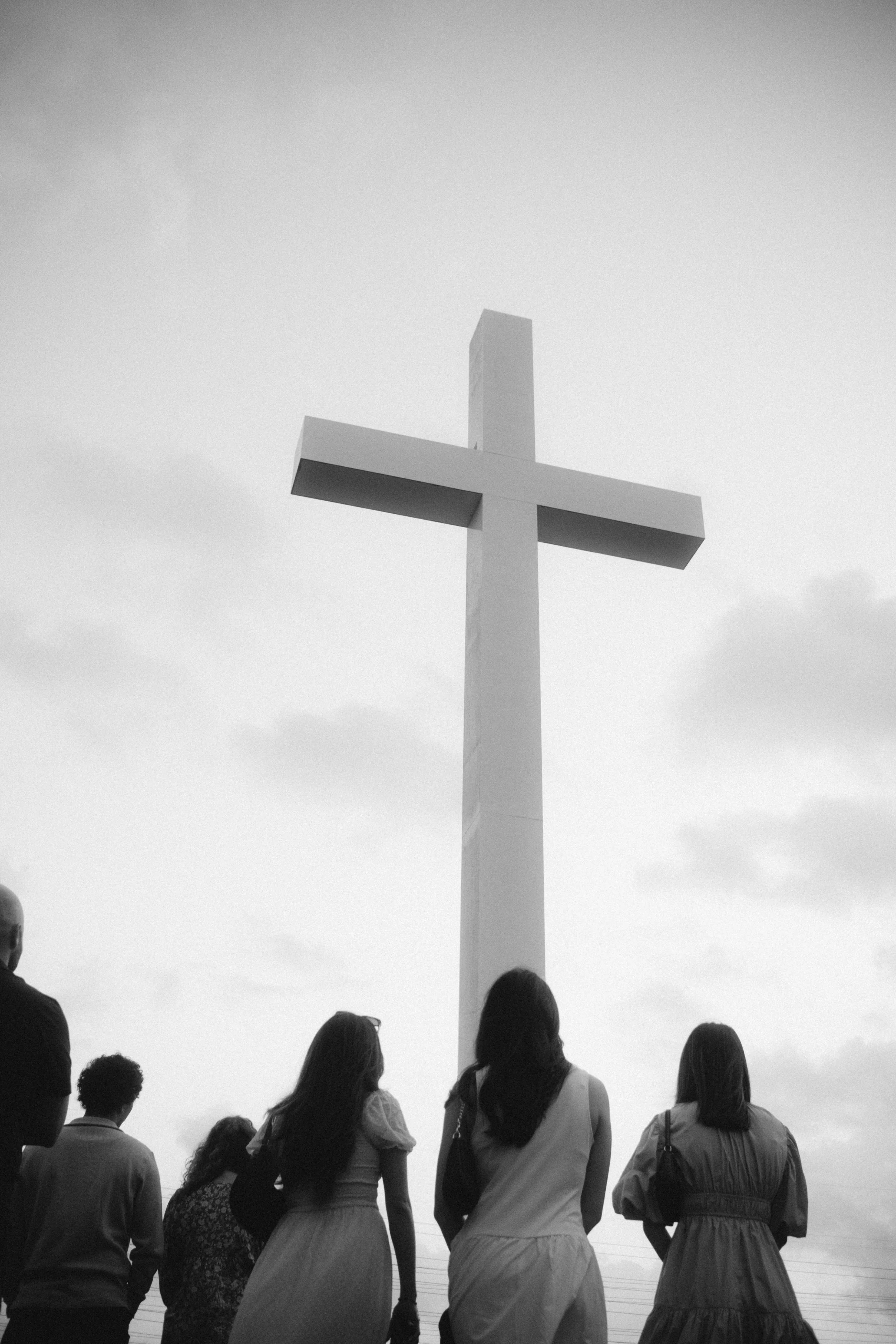 People standing in front of a large cross monument against a cloudy sky.