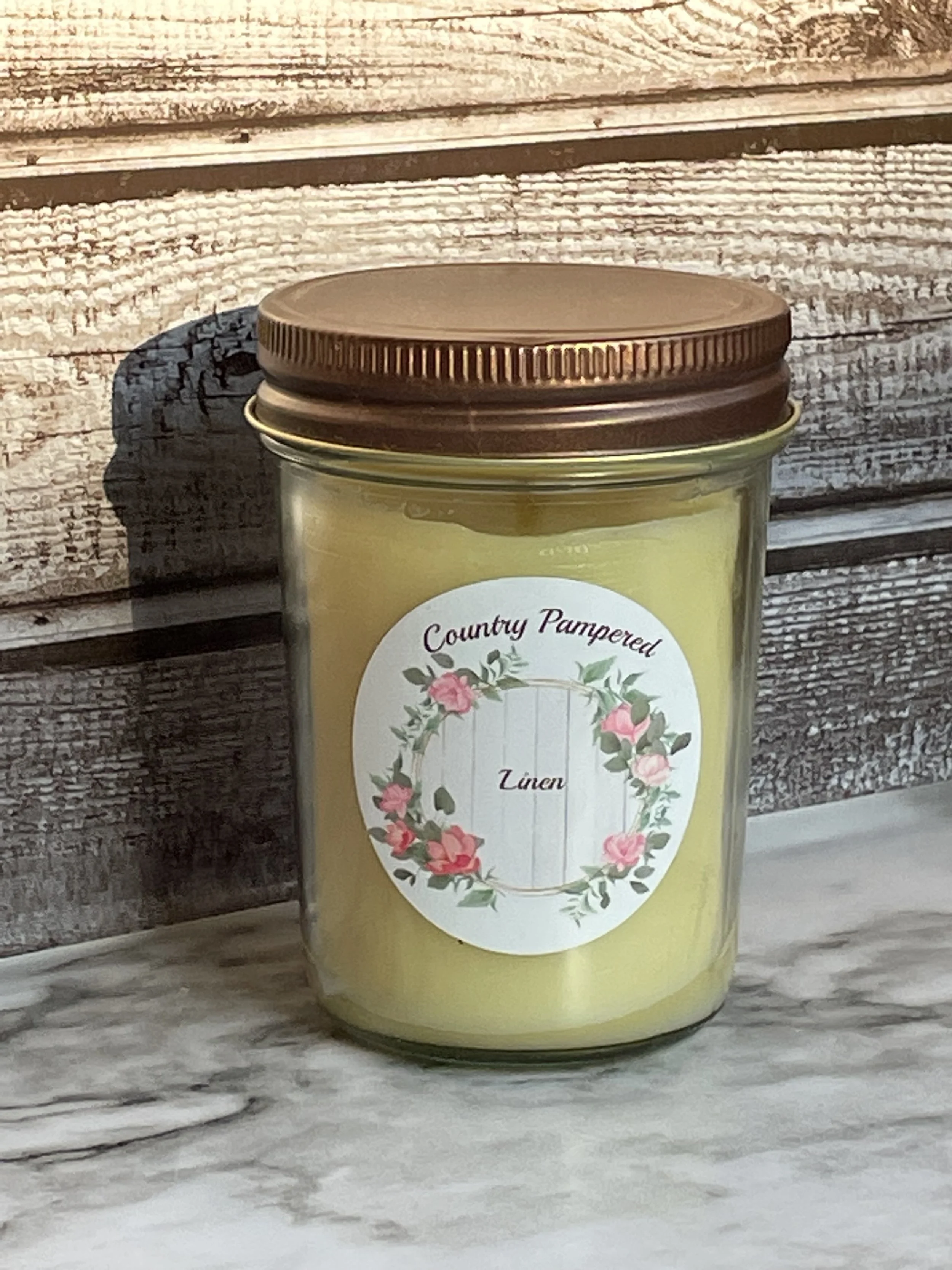 A glass jar of Linen scented candle labeled 'Country Pampered' with a floral wreath design on the front, placed on a wooden surface.