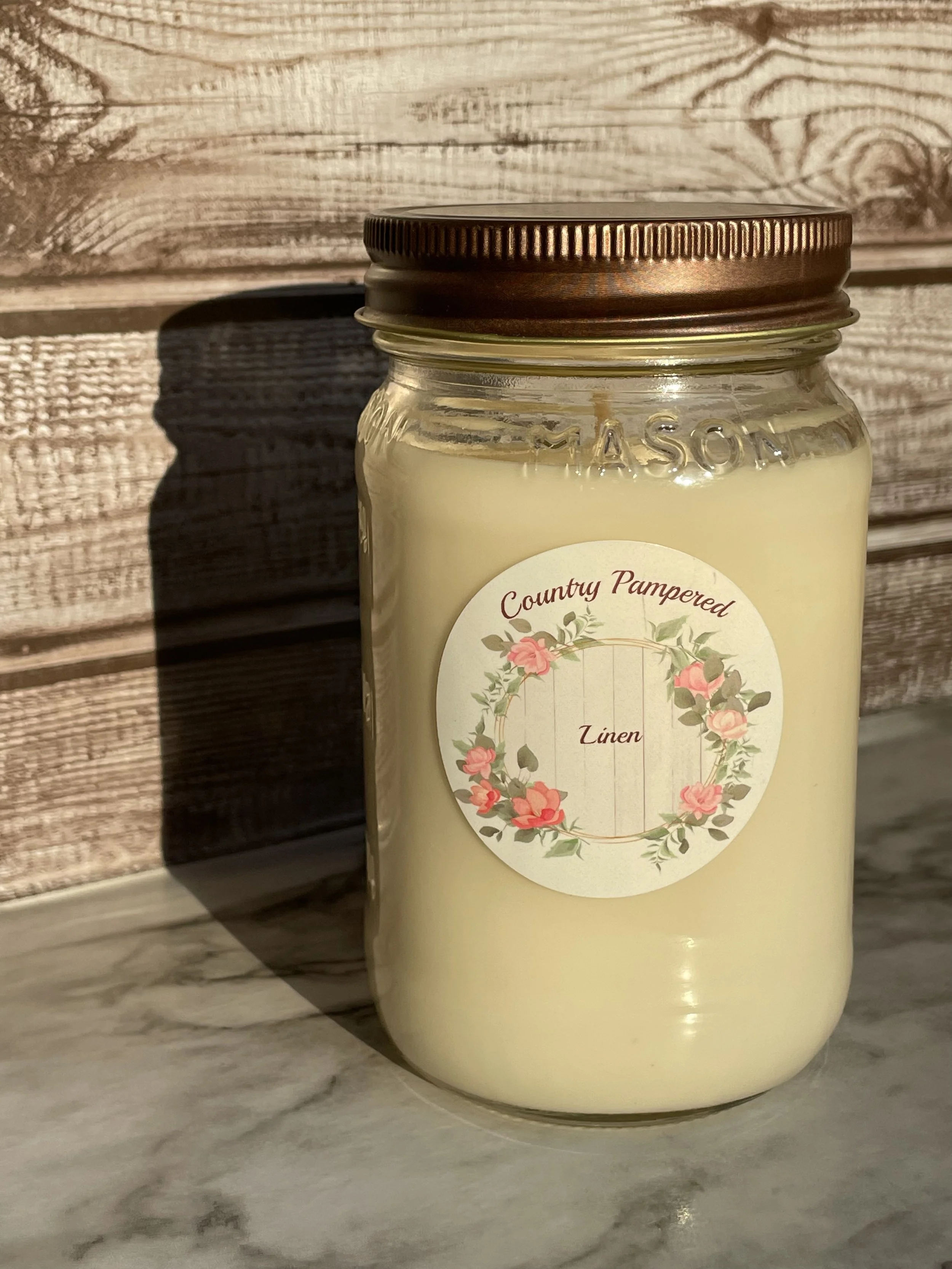 A glass jar of Linen scented candle labeled 'Country Pampered' with a floral wreath design on the front, placed on a wooden surface.