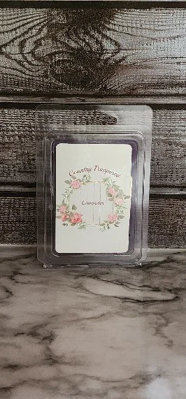 A container of Lavender scented wax melts labeled 'Country Pampered' with a floral wreath design on the front, placed on a wooden surface.