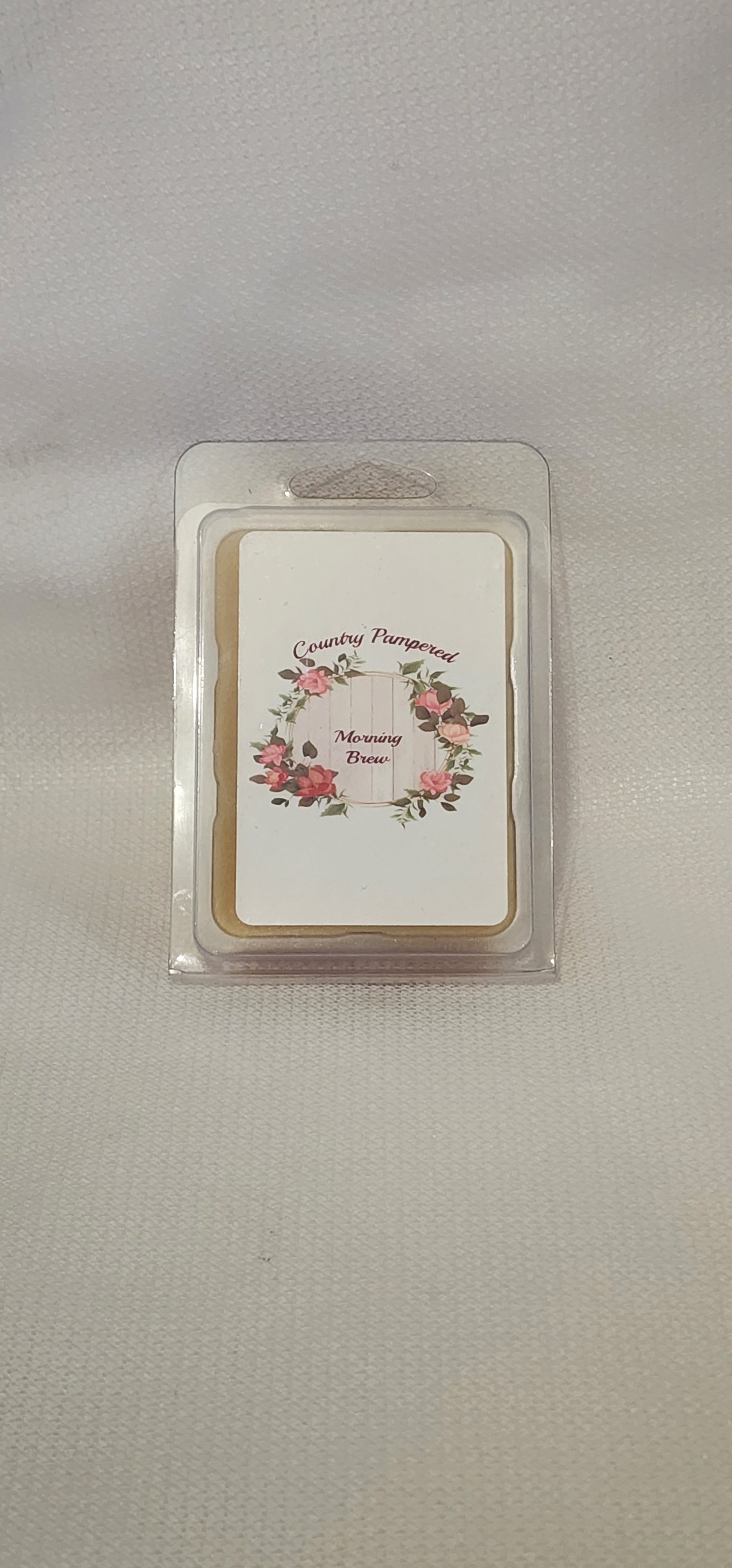 A container of Morning Brew scented wax melts labeled 'Country Pampered' with a floral wreath design on the front, placed on a wooden surface.