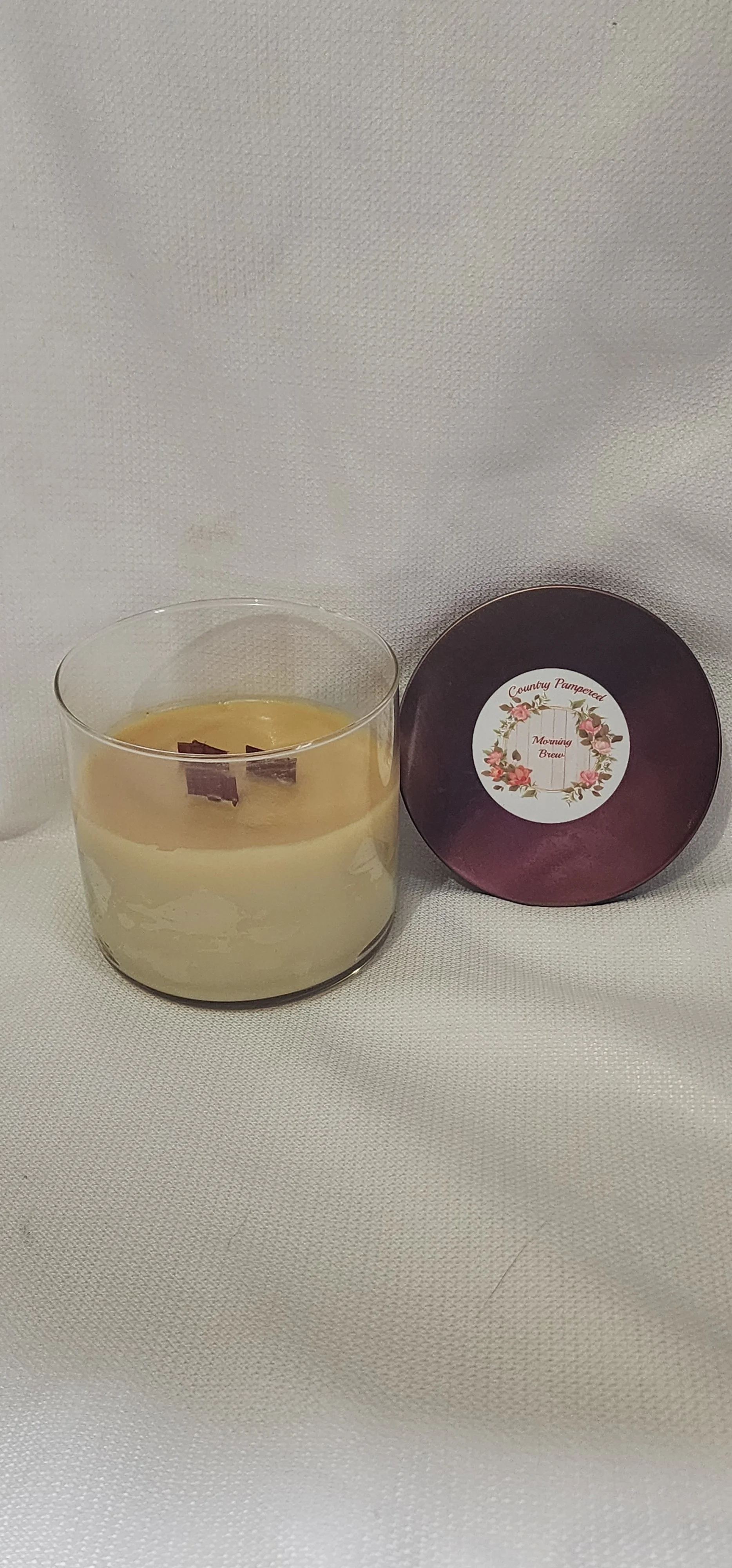 A glass jar of Morning Brew scented candle labeled 'Country Pampered' with a floral wreath design on the front, placed on a wooden surface.