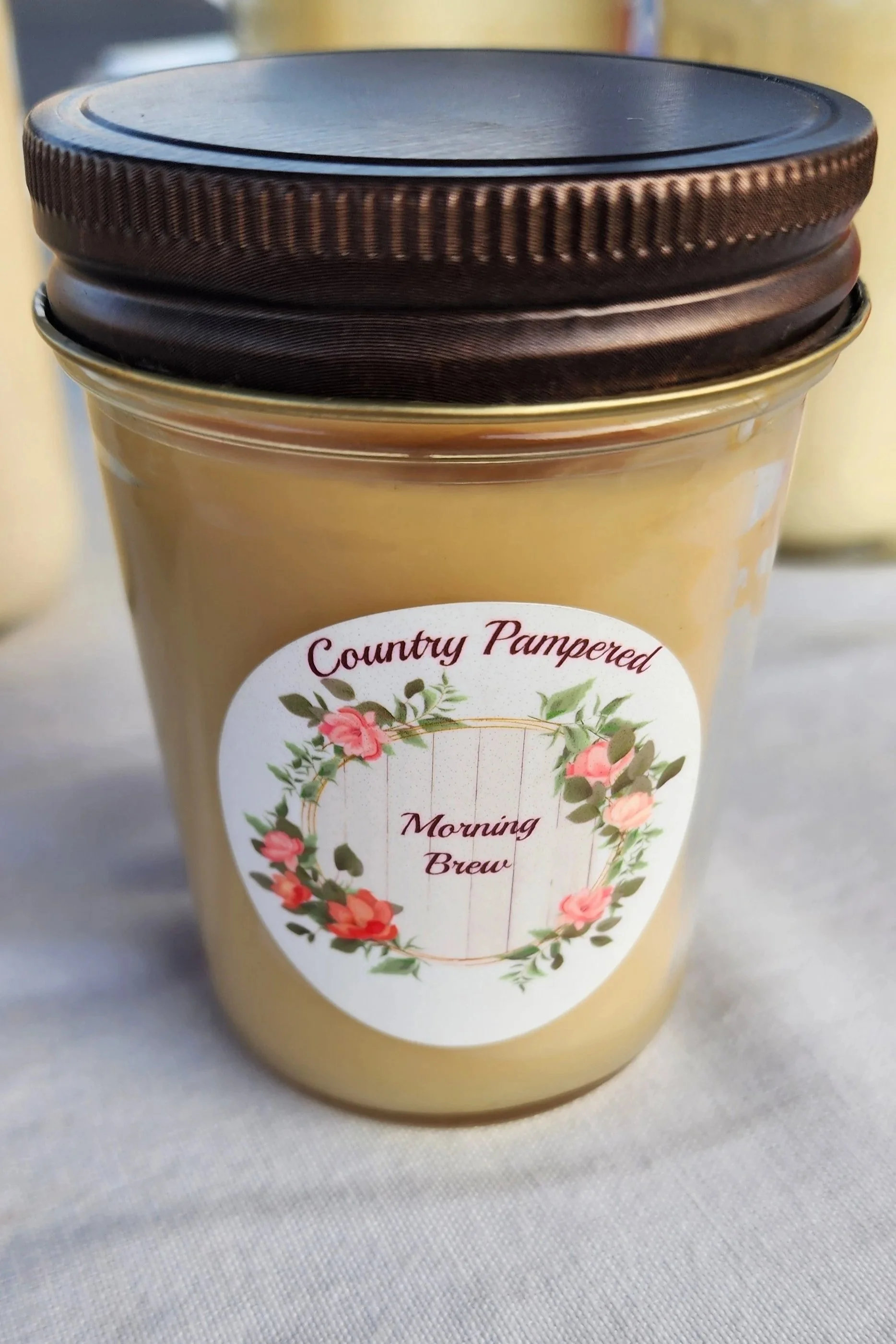 A glass jar of Morning Brew scented candle labeled 'Country Pampered' with a floral wreath design on the front, placed on a wooden surface.