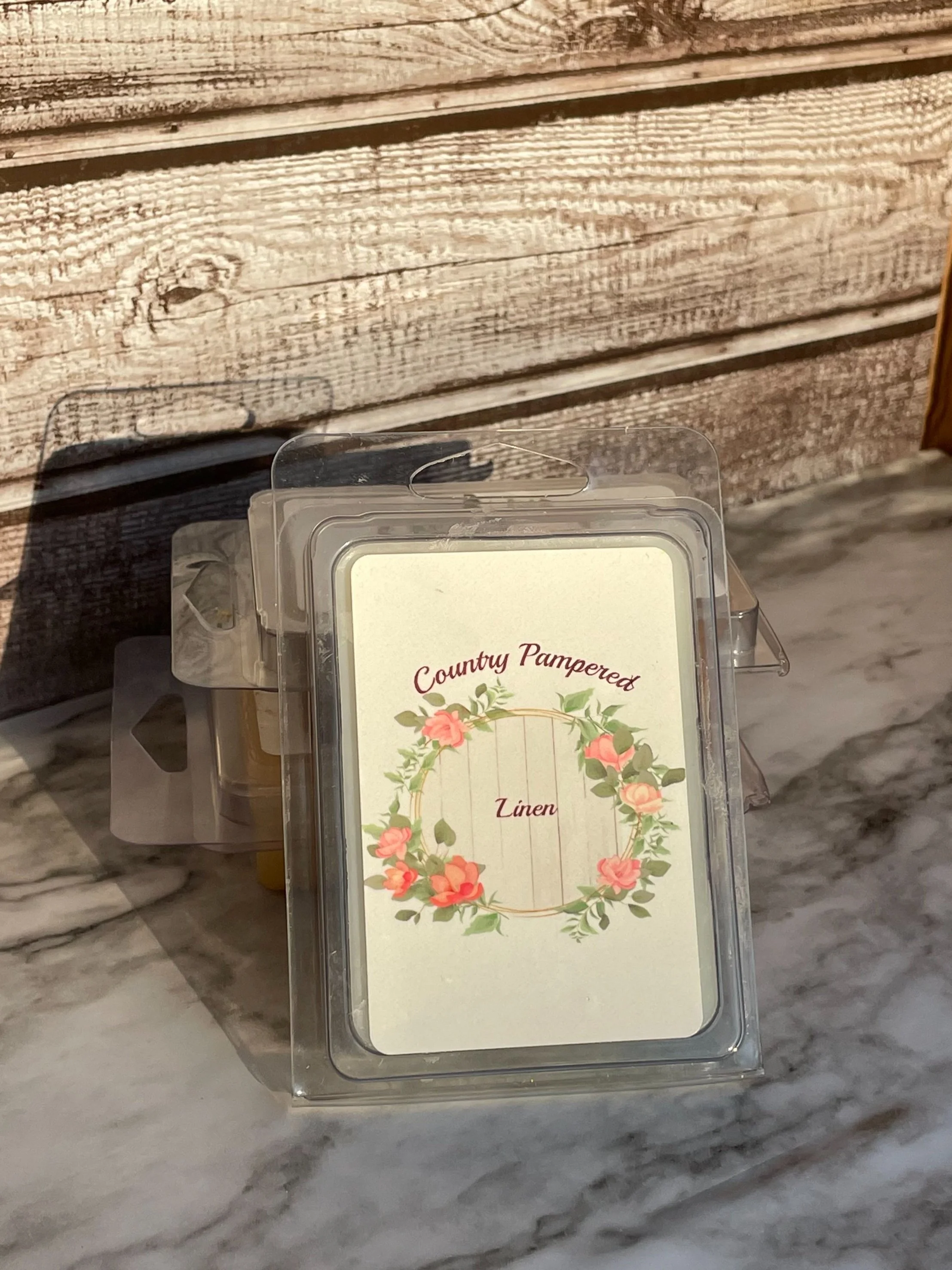 A container of Linen scented wax melts labeled 'Country Pampered' with a floral wreath design on the front, placed on a wooden surface.