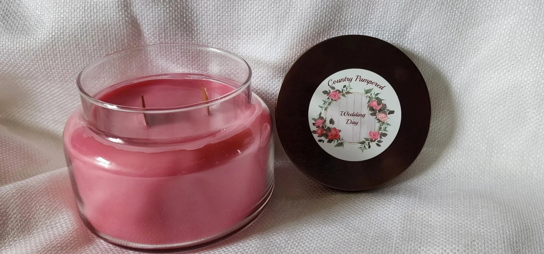 A glass jar of Wedding Day scented candle labeled 'Country Pampered' with a floral wreath design on the front, placed on a wooden surface.