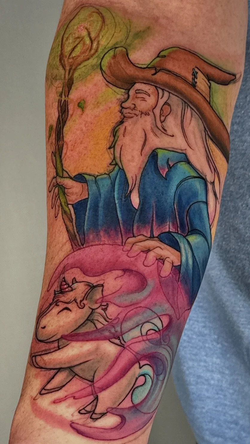 Colorful tattoo of a wizard with a long beard and hat holding a staff, with a unicorn in front of him, surrounded by pink and purple smoke.