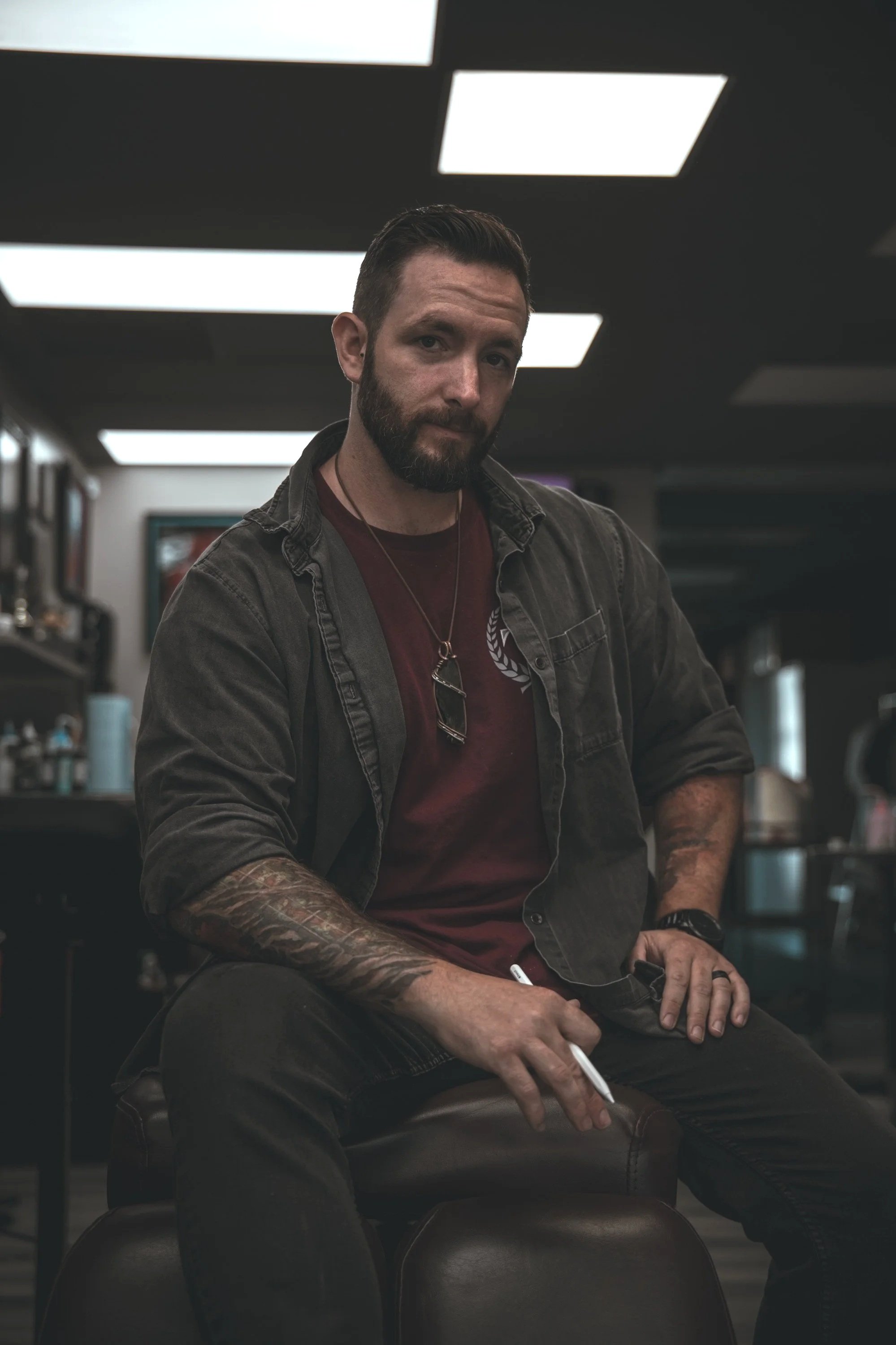 A man with dark hair, beard, and tattoos on his arm, wearing a dark jacket and red shirt, sitting in a dimly lit room, possibly a tattoo parlor or studio, holding a white pen or marker, with a serious expression on his face.
