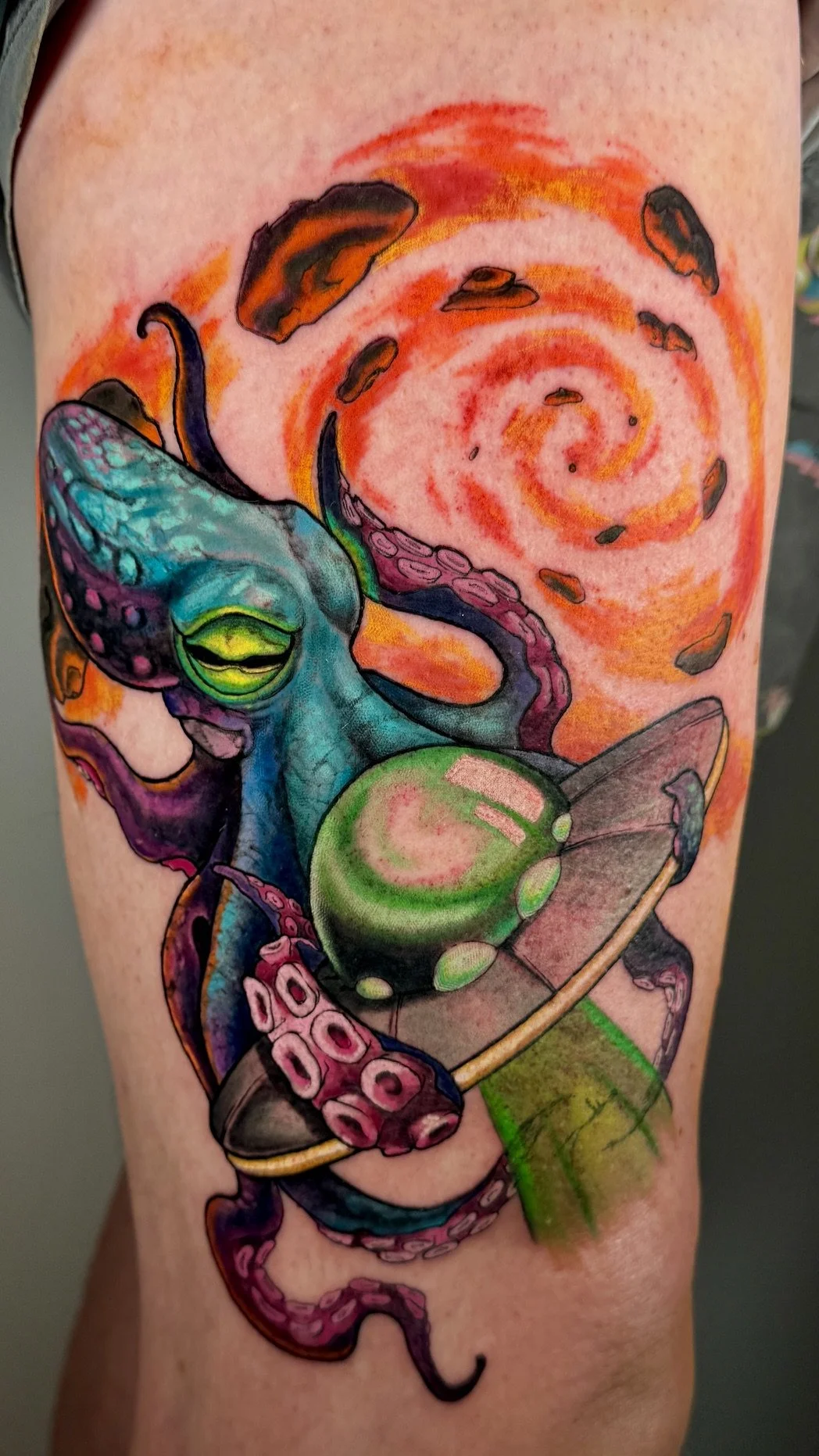 Colorful tattoo of a chameleon holding a green ball, with swirling red and orange background and scattered rocks.