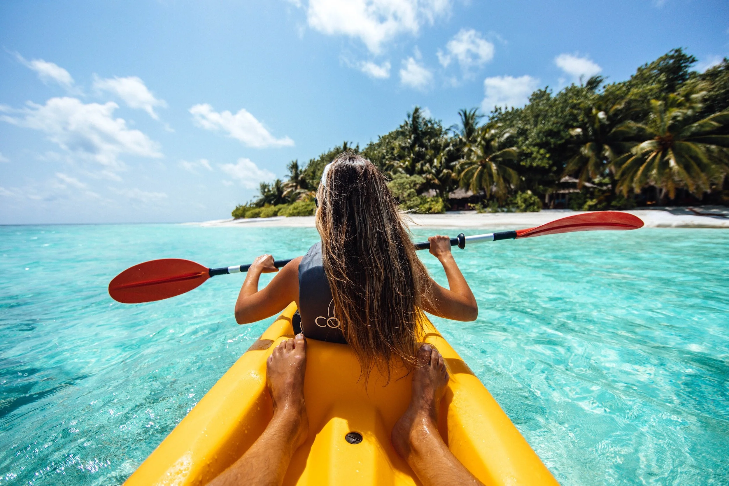 Person kayaking in turquoise water near a tropical island with palm trees, under a clear blue sky.