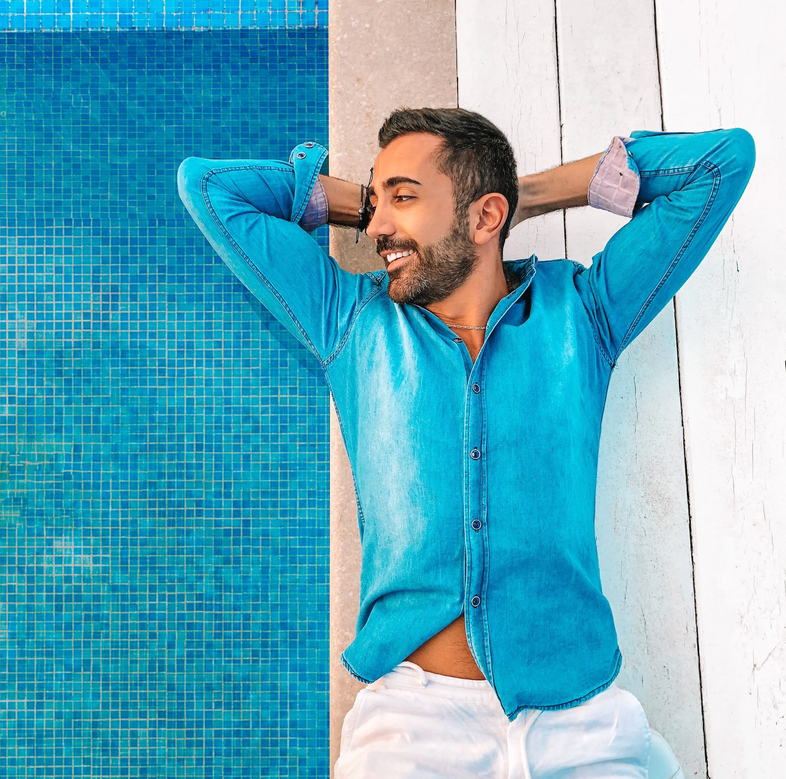 Man in denim shirt and white pants relaxing by a blue tiled swimming pool, smiling with hands behind head.