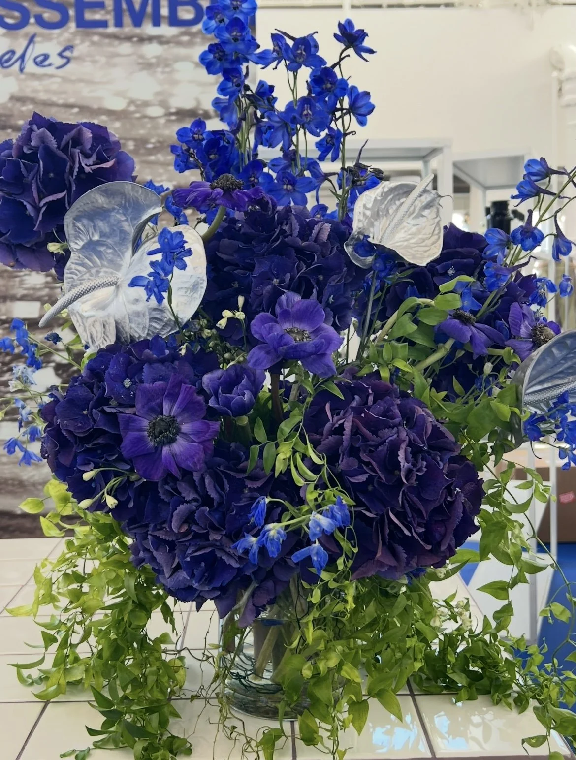 A floral arrangement featuring purple and blue flowers, including hydrangeas and delphiniums, accented with white butterfly decorations and green trailing foliage in a glass vase.
