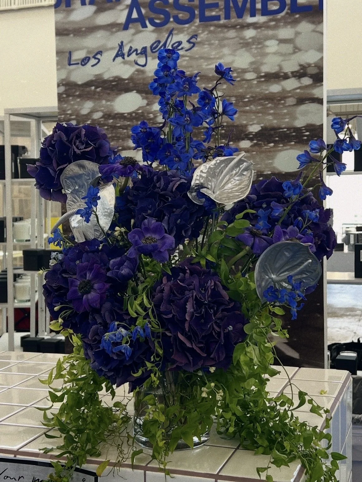 A flower arrangement with purple and blue flowers, including anthuriums, delphiniums, and petunias, with trailing green vines in a glass vase, displayed on a tiled surface.