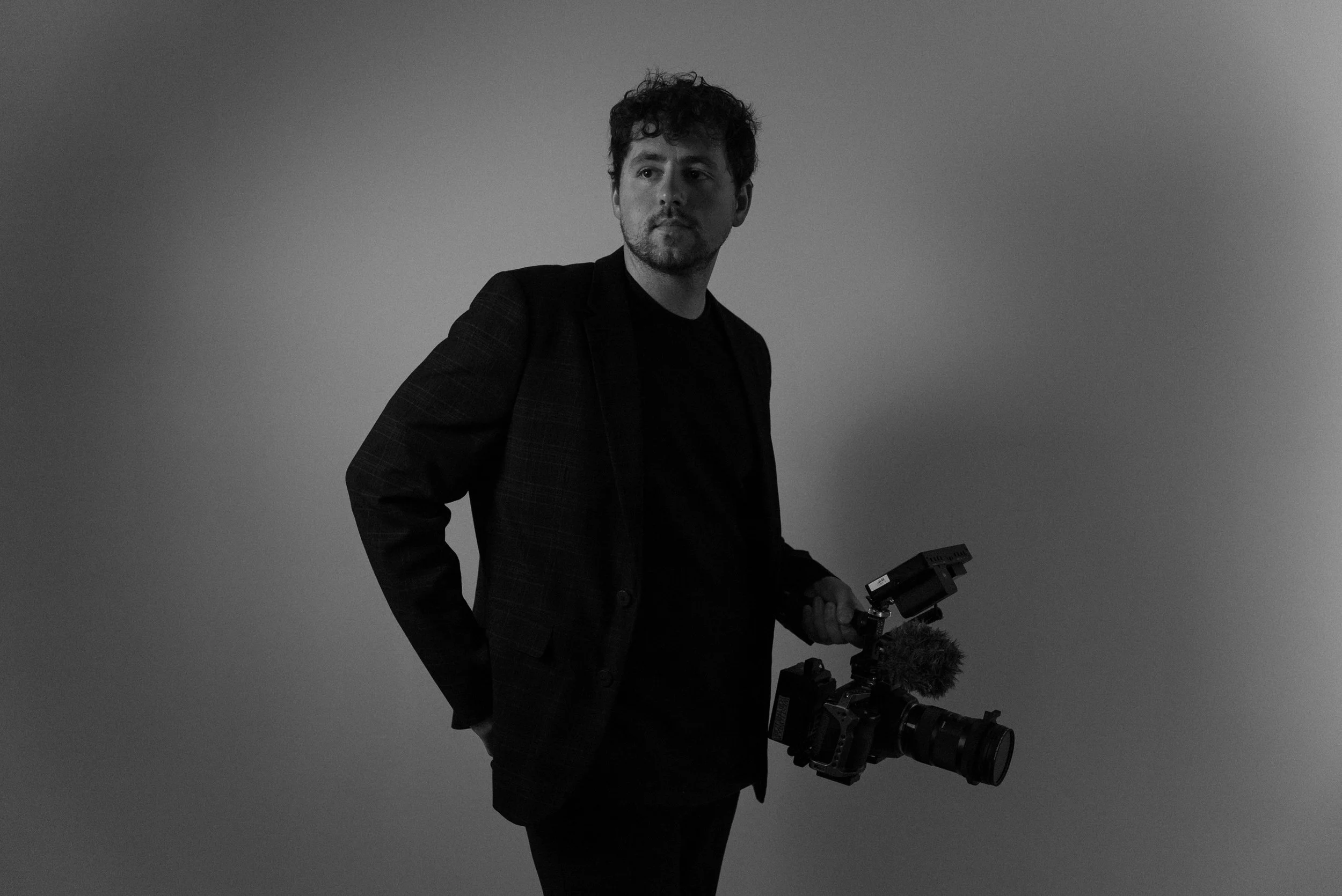 Black and white photo of a man holding a professional video camera, wearing a dark blazer, standing against a plain background.