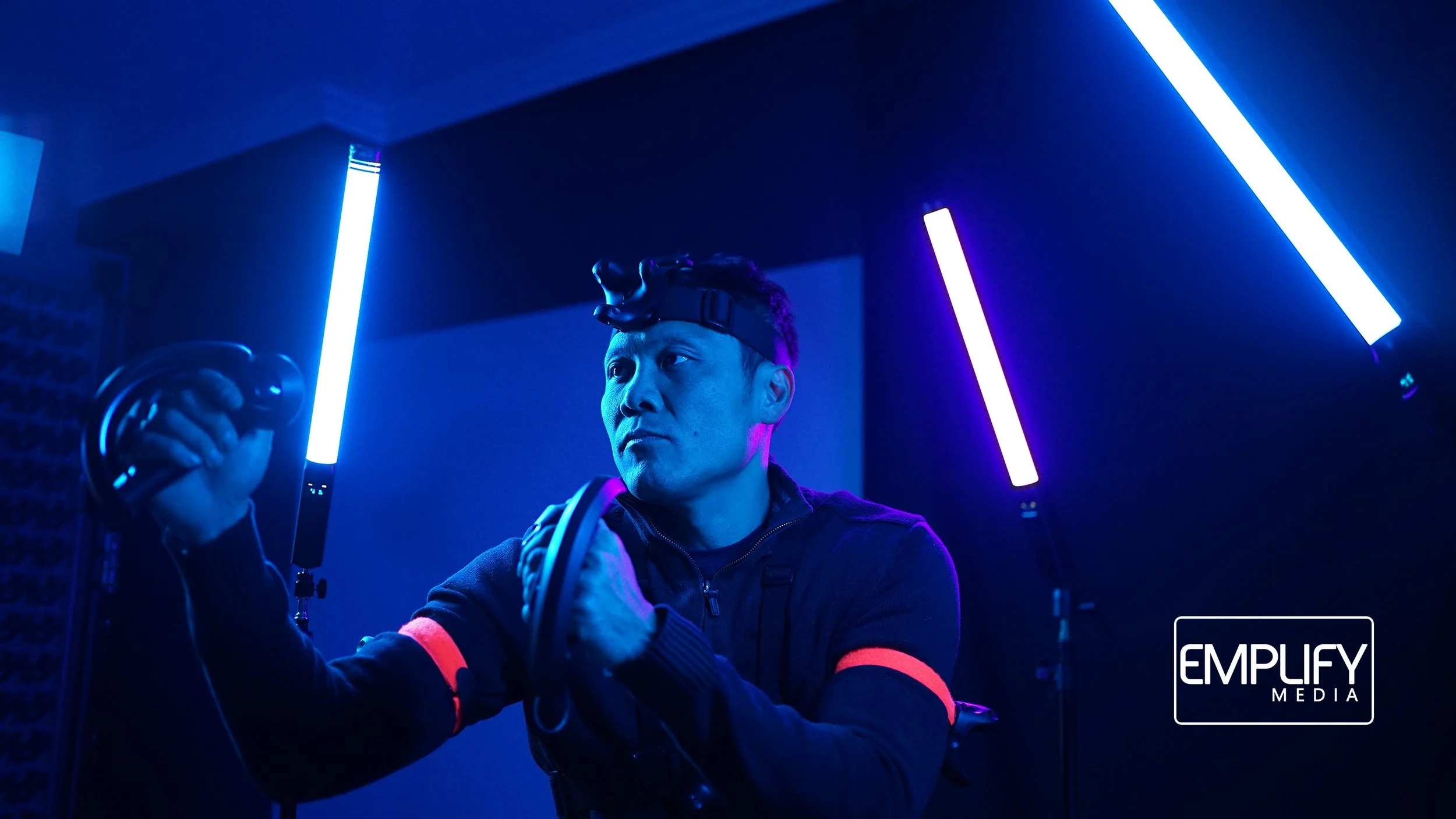 A man in a dark room with blue and purple neon lights, holding a VR headset and a controller, looking serious.