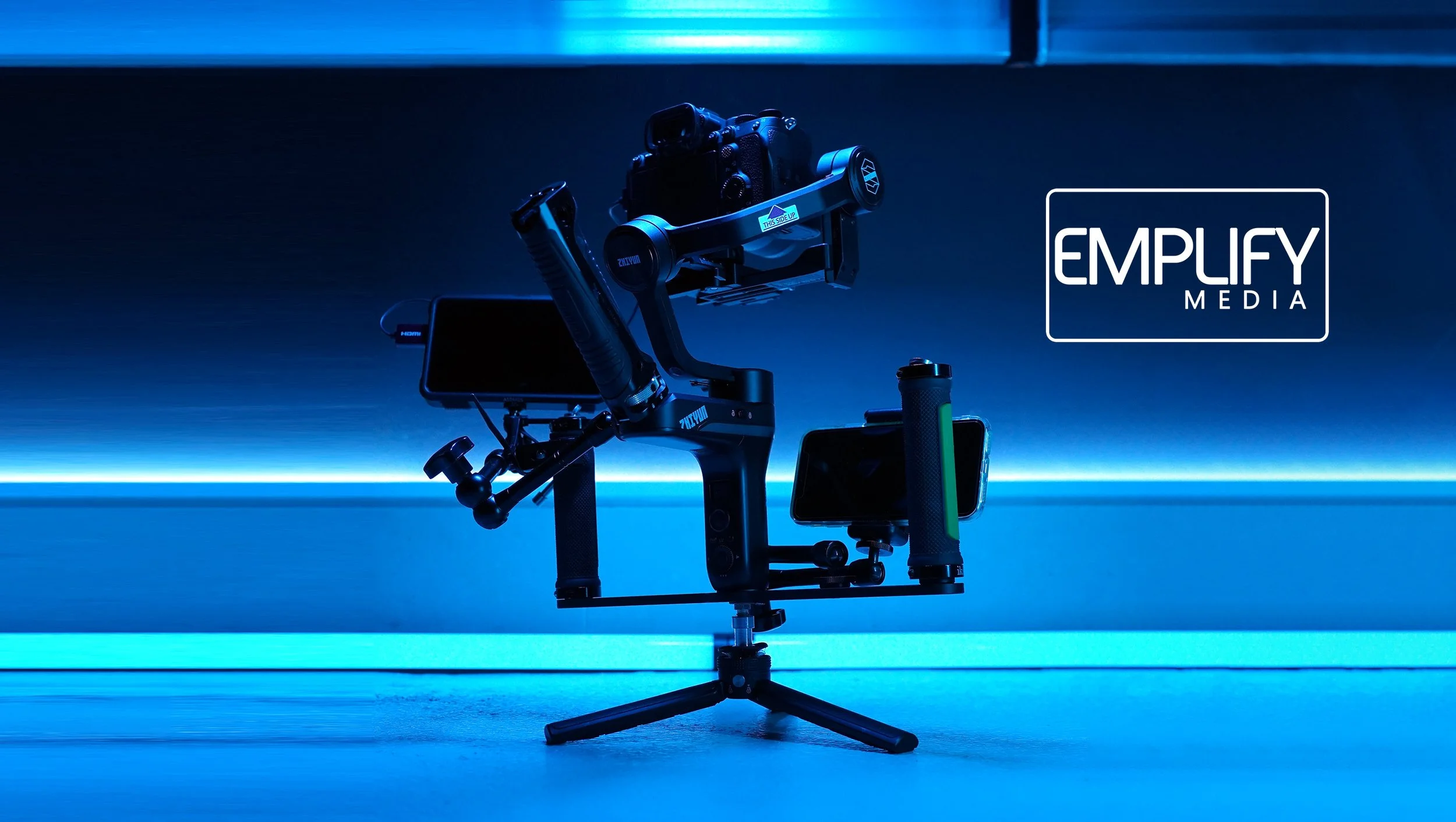 Camera mounted on a gimbal with two attached smartphones, set against a blue-lit background, with "EMPLYFY MEDIA" logo on the right.