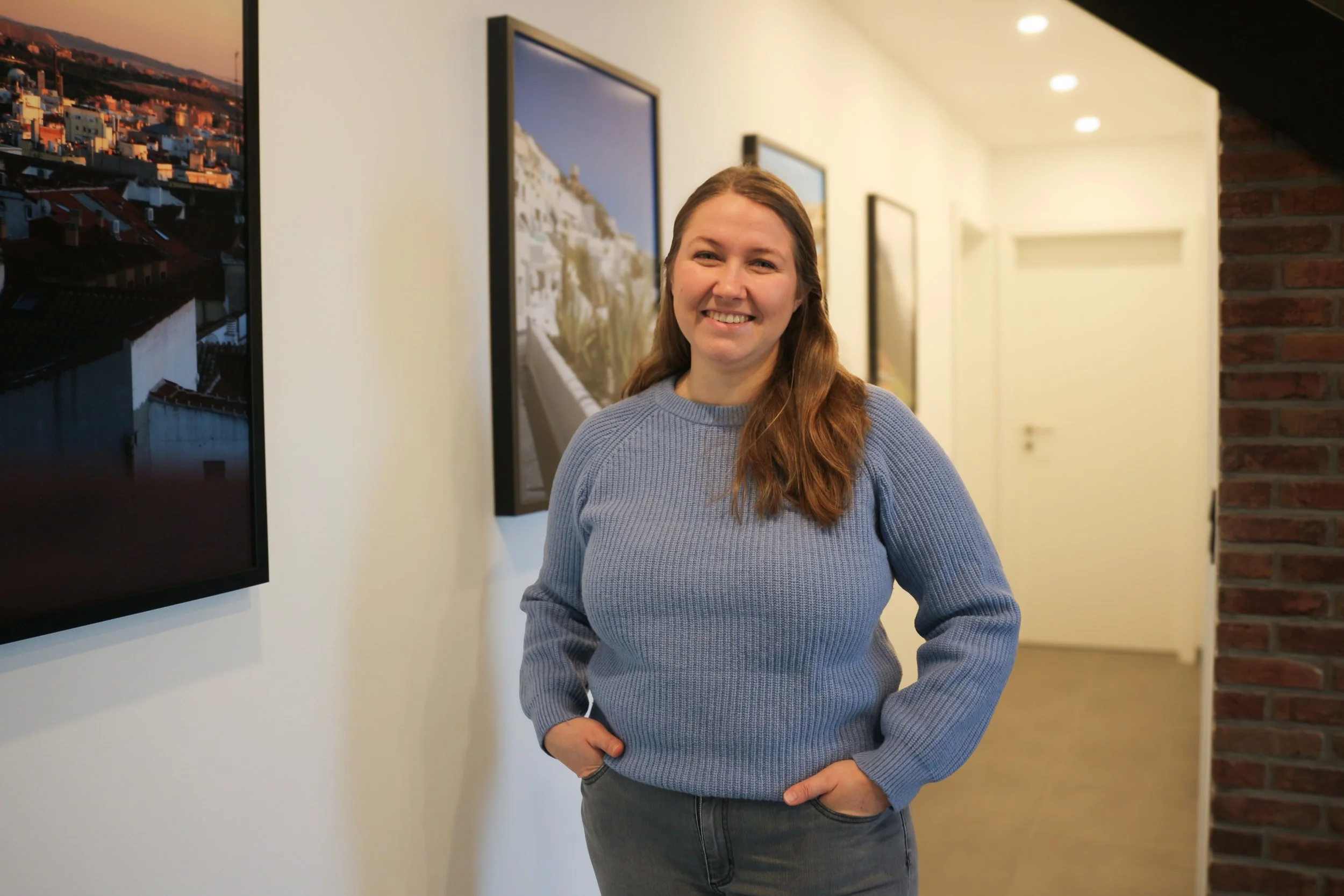 Lächelnde Frau mit blonden Haaren in blauer Strickjacke in einer Galerie mit Kunstfotografien an der Wand.
