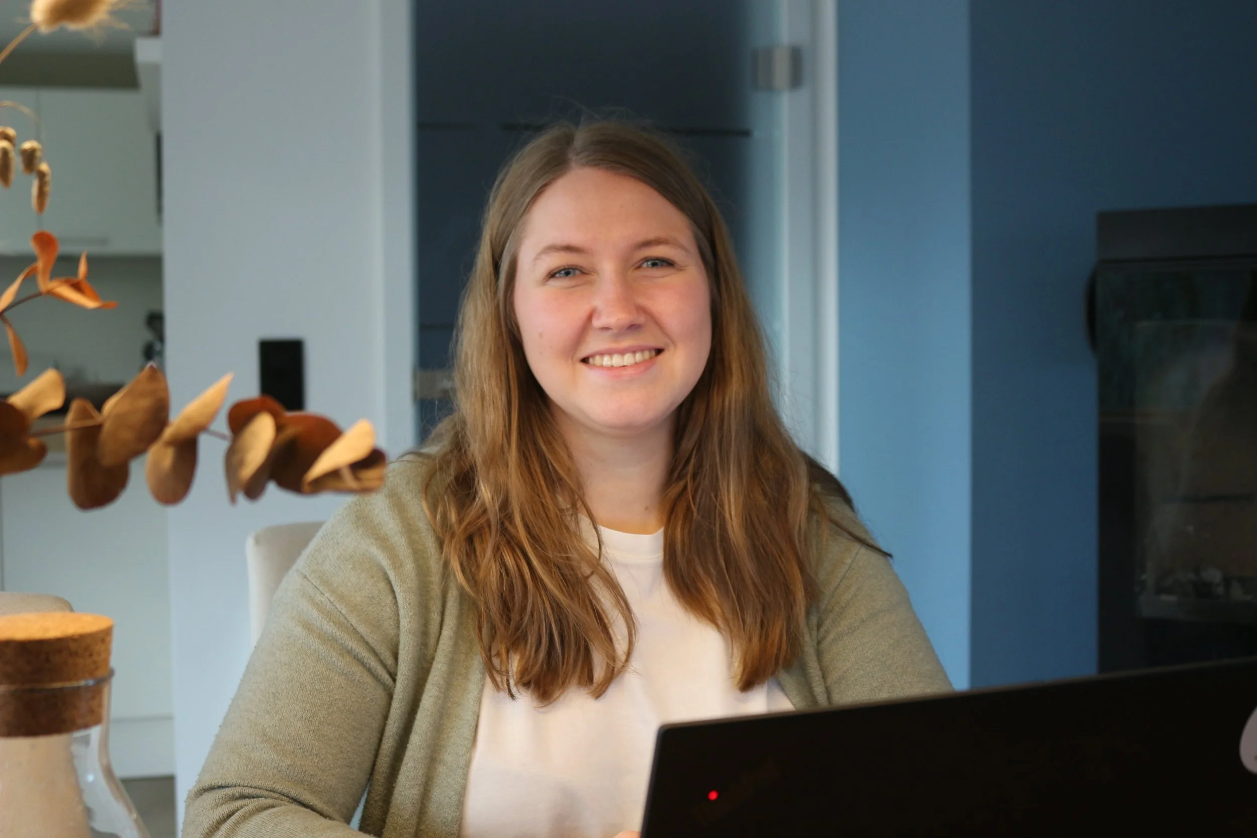 Junge Frau mit langen blonden Haaren, lächelnd, sitzt an einem Tisch mit Laptop in einer modernen Küche