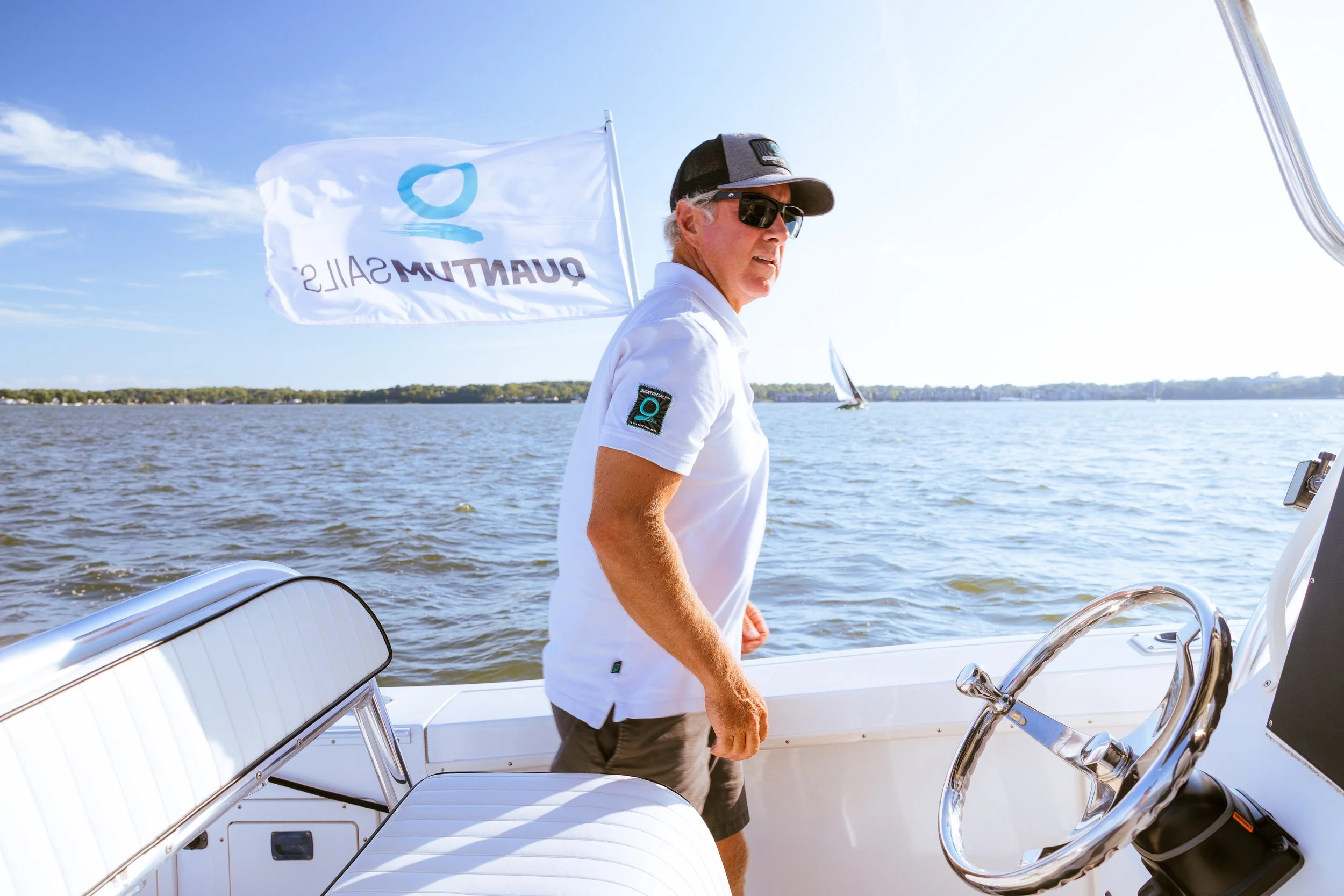 Man with gray hair wearing sunglasses, a white polo shirt, and a black cap, standing on a boat, steering with an ocean and a sailboat in the background, and a flag with 'Quantum Sails' logo in the wind.