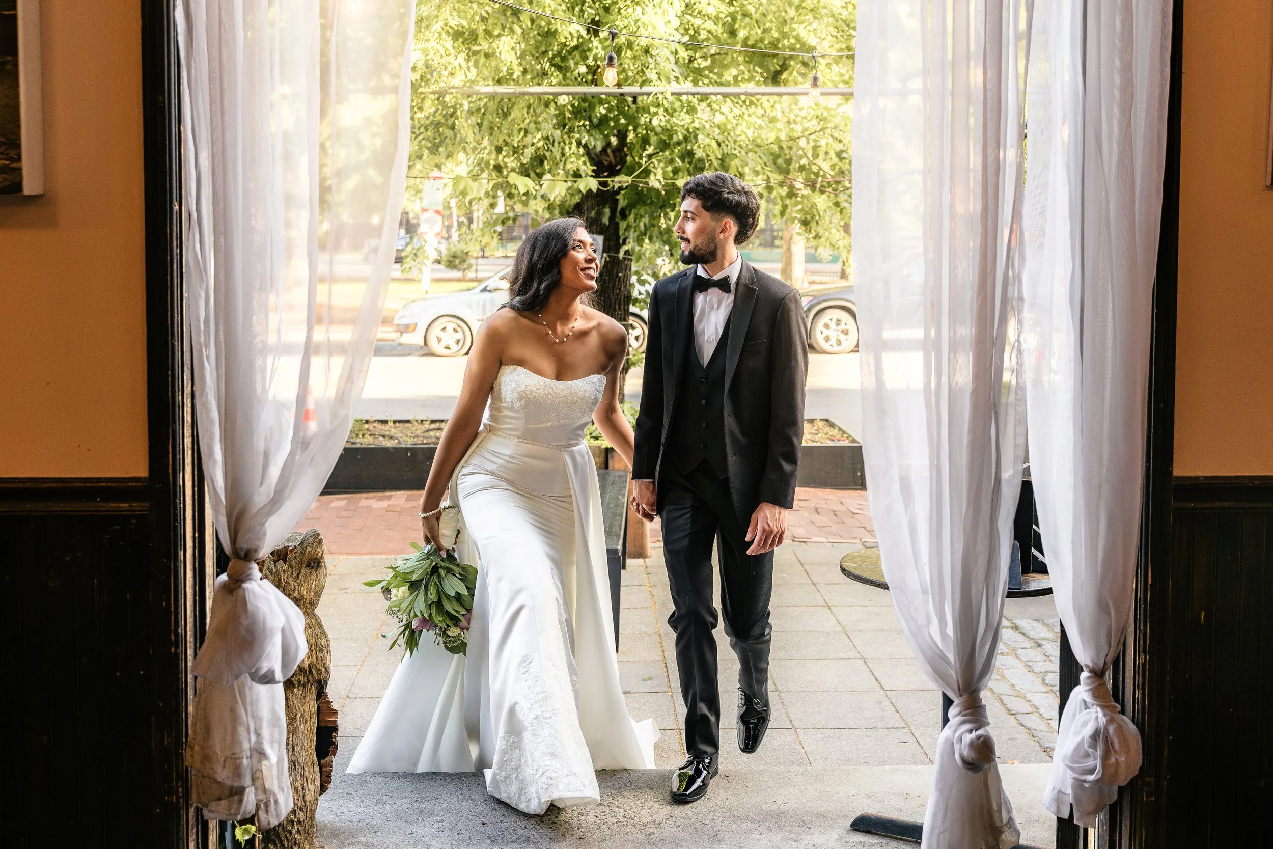 A bride and groom walk arm in arm outside a venue, framed by white curtains, with sunlight filtering through trees in the background.