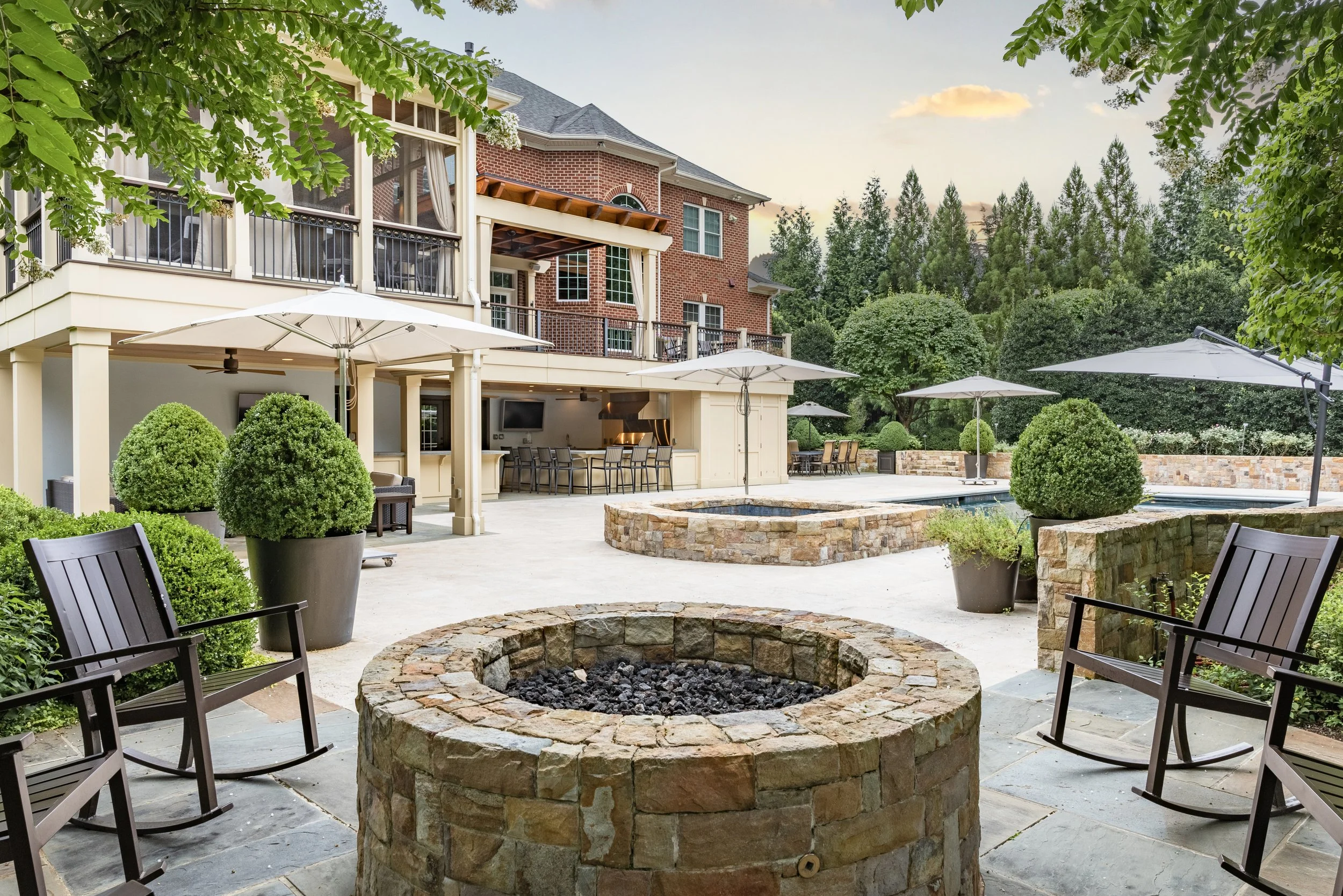 An outdoor patio area with a central fire pit, surrounded by chairs and potted shrubs, adjacent to a building with a balcony and bar area, under umbrellas and with a backdrop of trees.