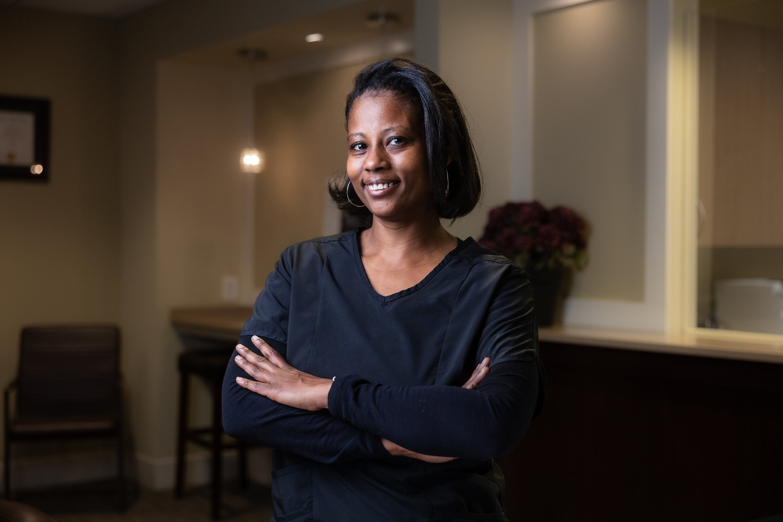 Woman with shoulder-length black hair smiling, wearing a black top with arms crossed, standing indoors with warm lighting and blurred background.
