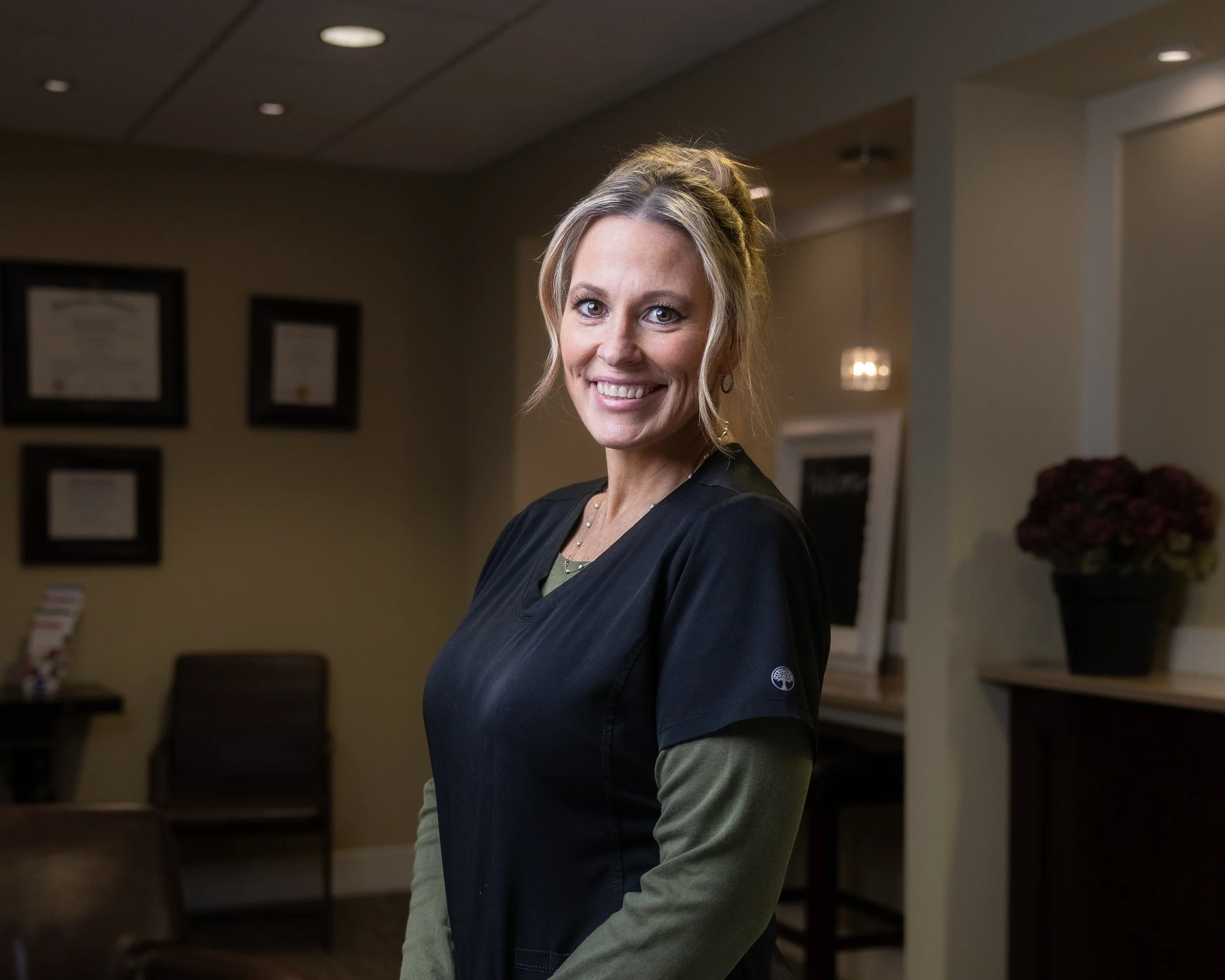A smiling woman in medical scrubs standing in a waiting area of a medical office or clinic, with framed certificates on the wall and a potted plant in the background.